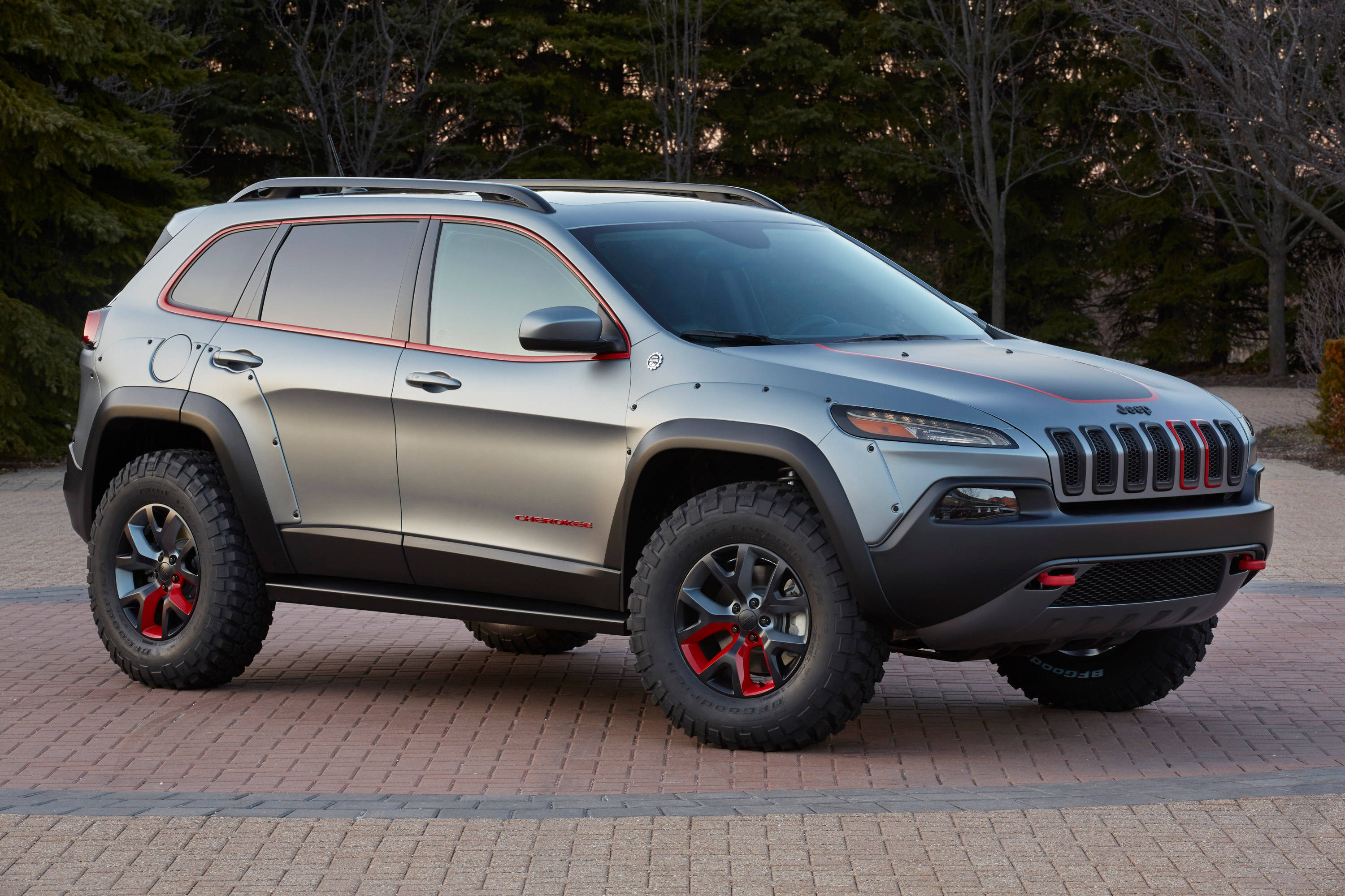 Jeep Cherokee 2015 Trailhawk 壁紙 — 一輛銀色Jeep Cherokee，帶有紅色點綴和堅固輪胎，停在磚砌車道上，周圍樹木環繞。