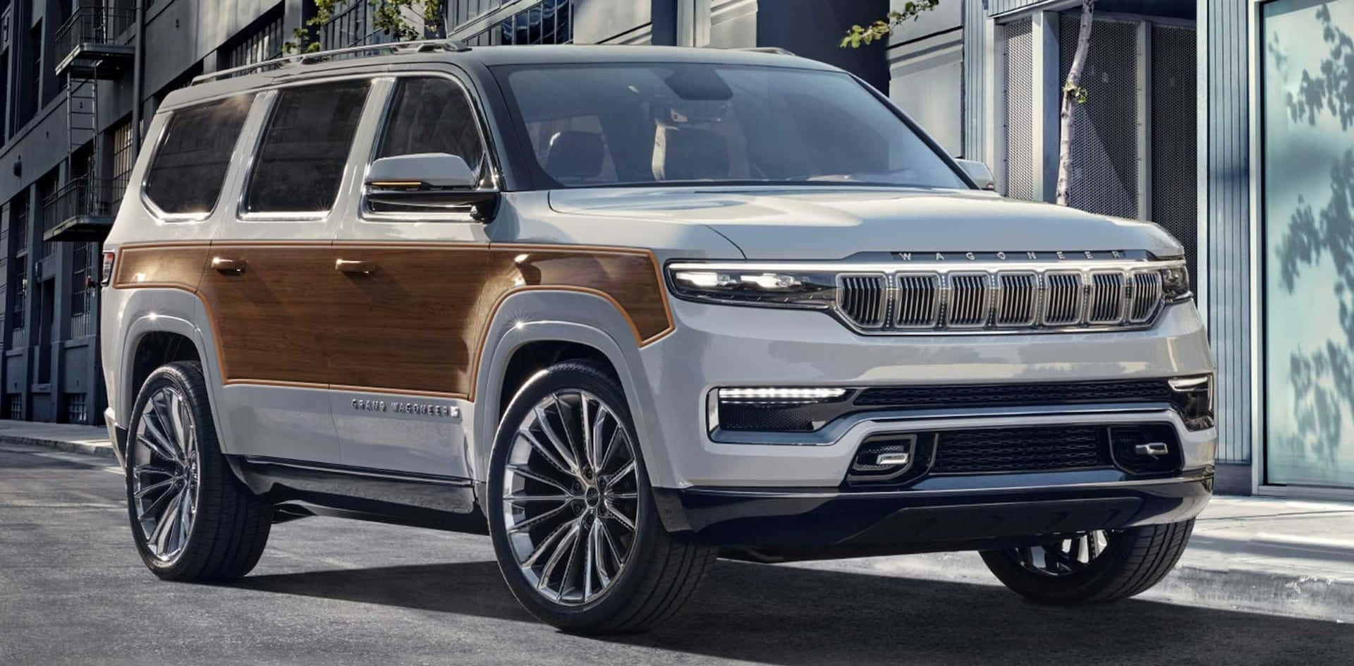 Jeep Wagoneer 桌布 壁紙 — 一輛銀色Jeep Grand Wagoneer SUV，帶有木紋鑲板，停在現代建築旁邊的城市街道上。
