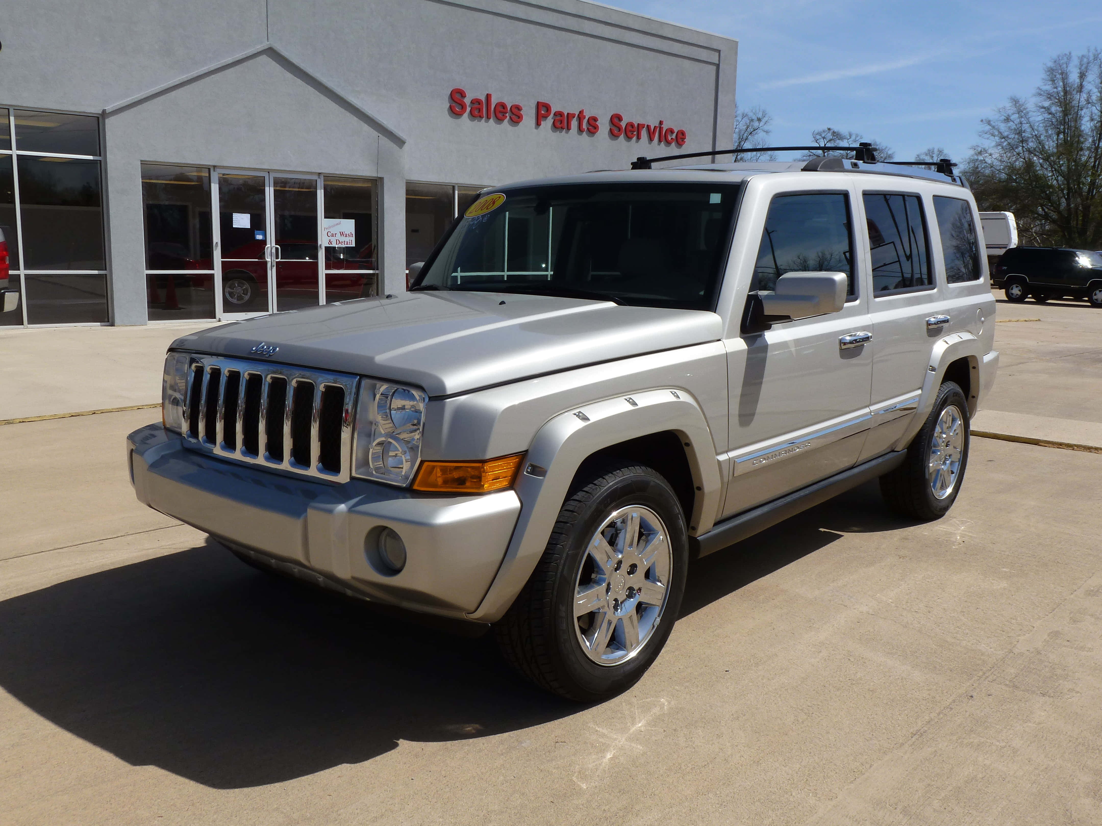 Jeep Commander 桌布 壁紙 — 一輛銀色Jeep Liberty SUV停泊在汽車經銷商前，背景是晴朗的藍天和樹木。