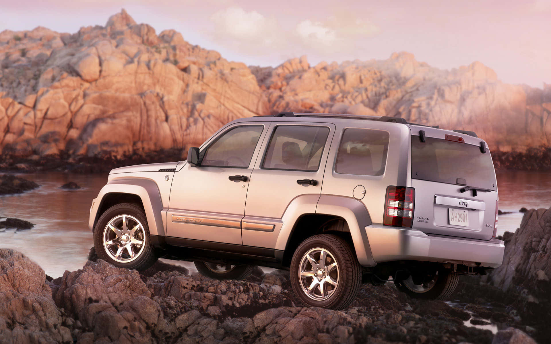 Jeep Liberty 桌布 壁紙 — 一輛銀色Jeep Liberty在日落時分穿越崎嶇的海岸岩石，背景是溫暖的橙色山脈。