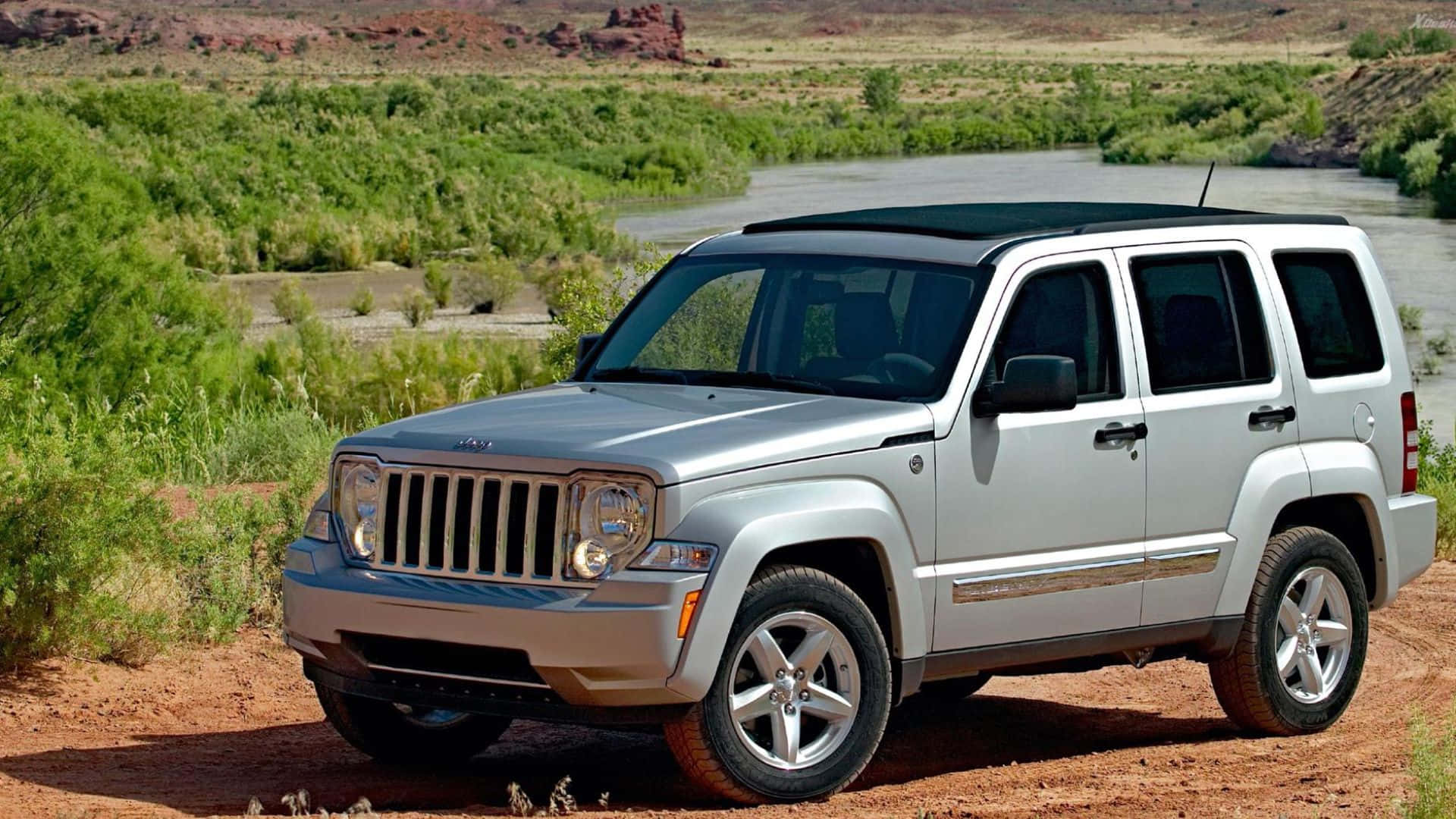 Jeep Liberty 桌布 壁紙 — 一輛銀色Jeep Liberty停泊在河邊的泥路上，周圍是鬱鬱蔥蔥的綠色沙漠景觀。