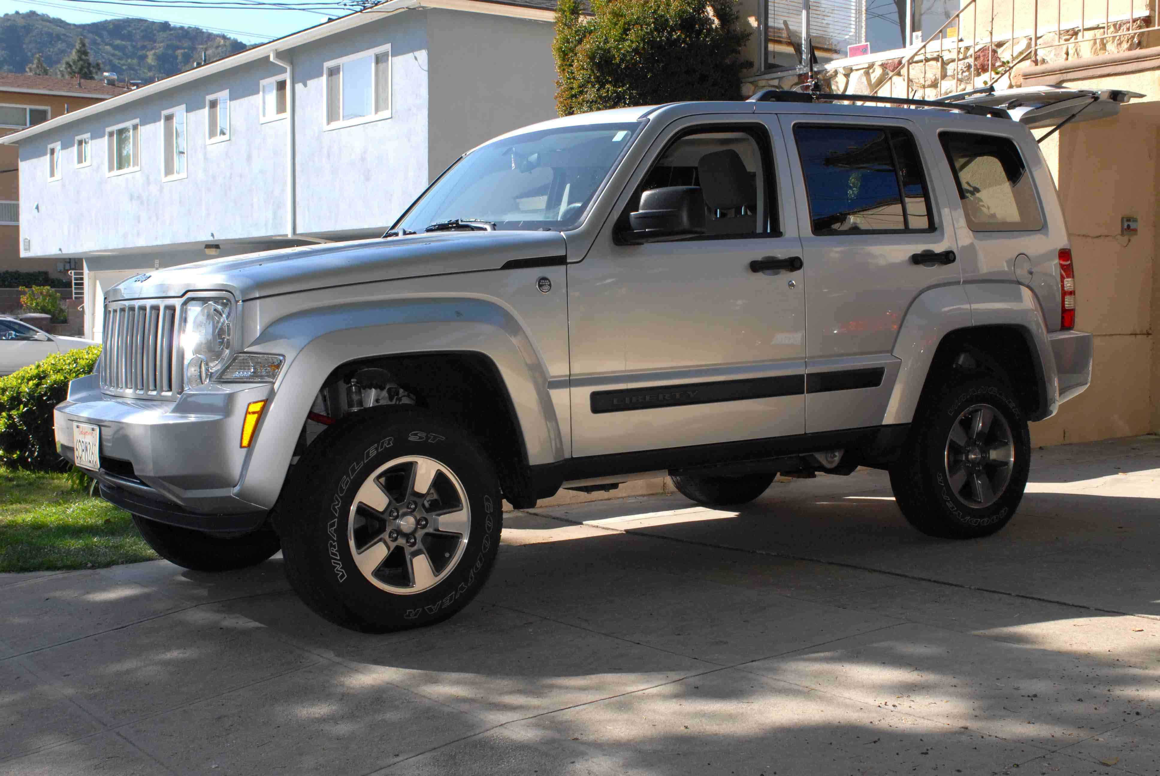 Jeep Liberty 桌布 壁紙 — 一輛銀色Jeep Liberty SUV停泊在車道上，背景是藍色房屋和綠色灌木叢，在明亮的陽光下。