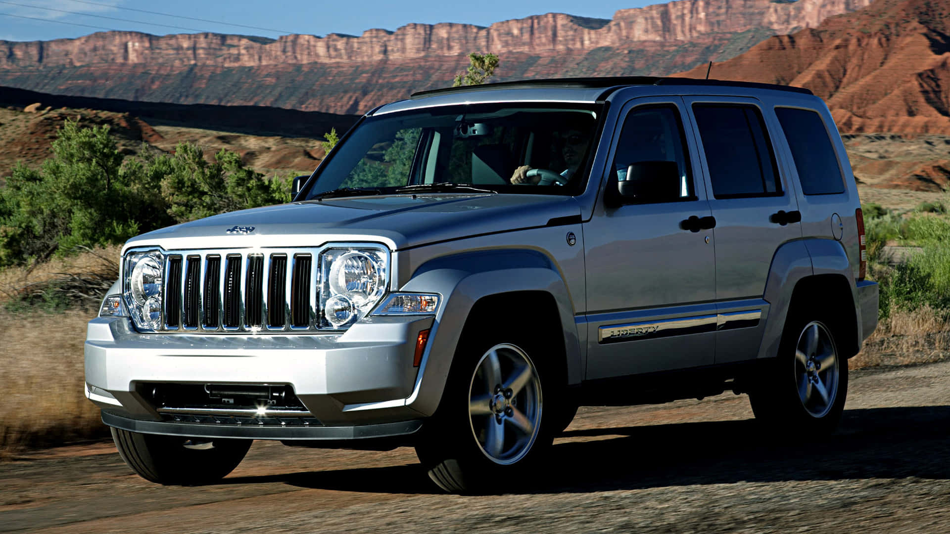 Jeep Liberty 桌布 壁紙 — 一輛銀色Jeep Liberty SUV停泊在泥路上，背景是紅色岩石峽谷懸崖，天空清澈湛藍。