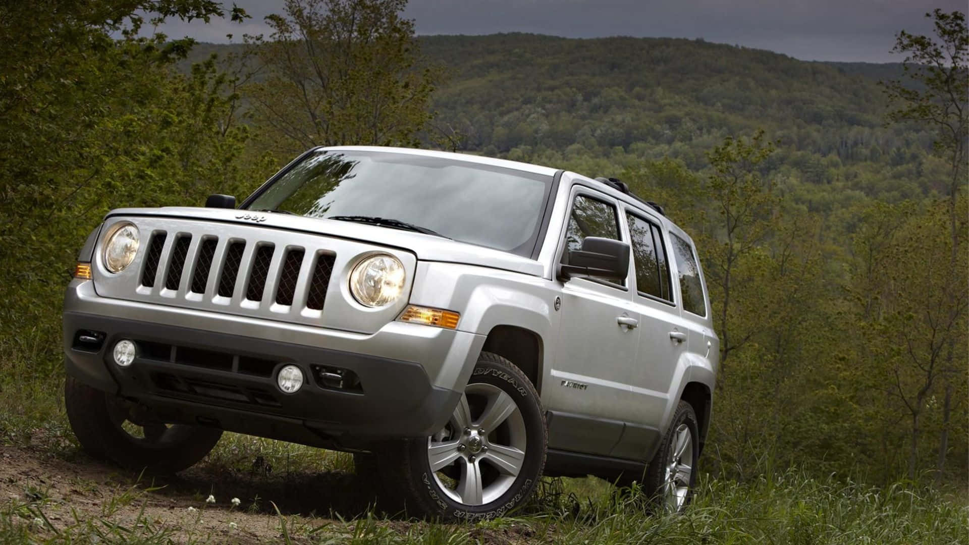 Jeep Patriot 桌布 壁紙 — 一輛銀色Jeep Patriot SUV停泊在草地小徑上，周圍是茂密的綠色森林和起伏的山丘，天空多雲。