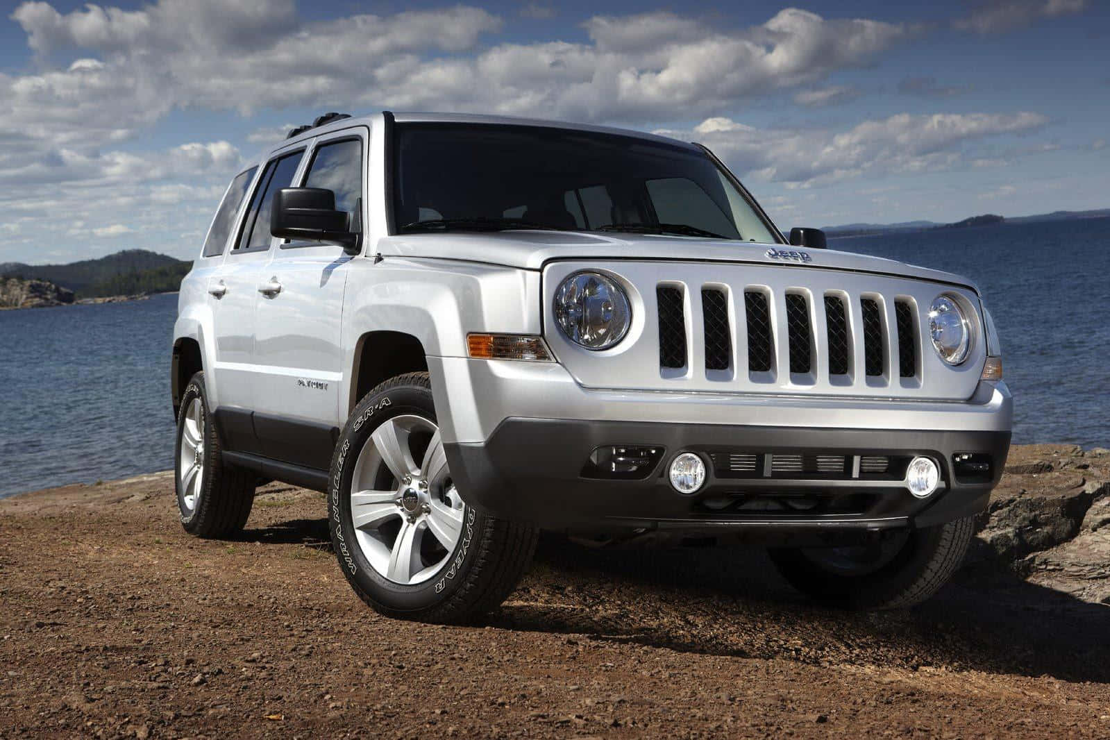 Jeep Patriot 桌布 壁紙 — 一輛銀色Jeep Patriot SUV停泊在岩石湖岸上，天空部分多雲，遠處有山丘。
