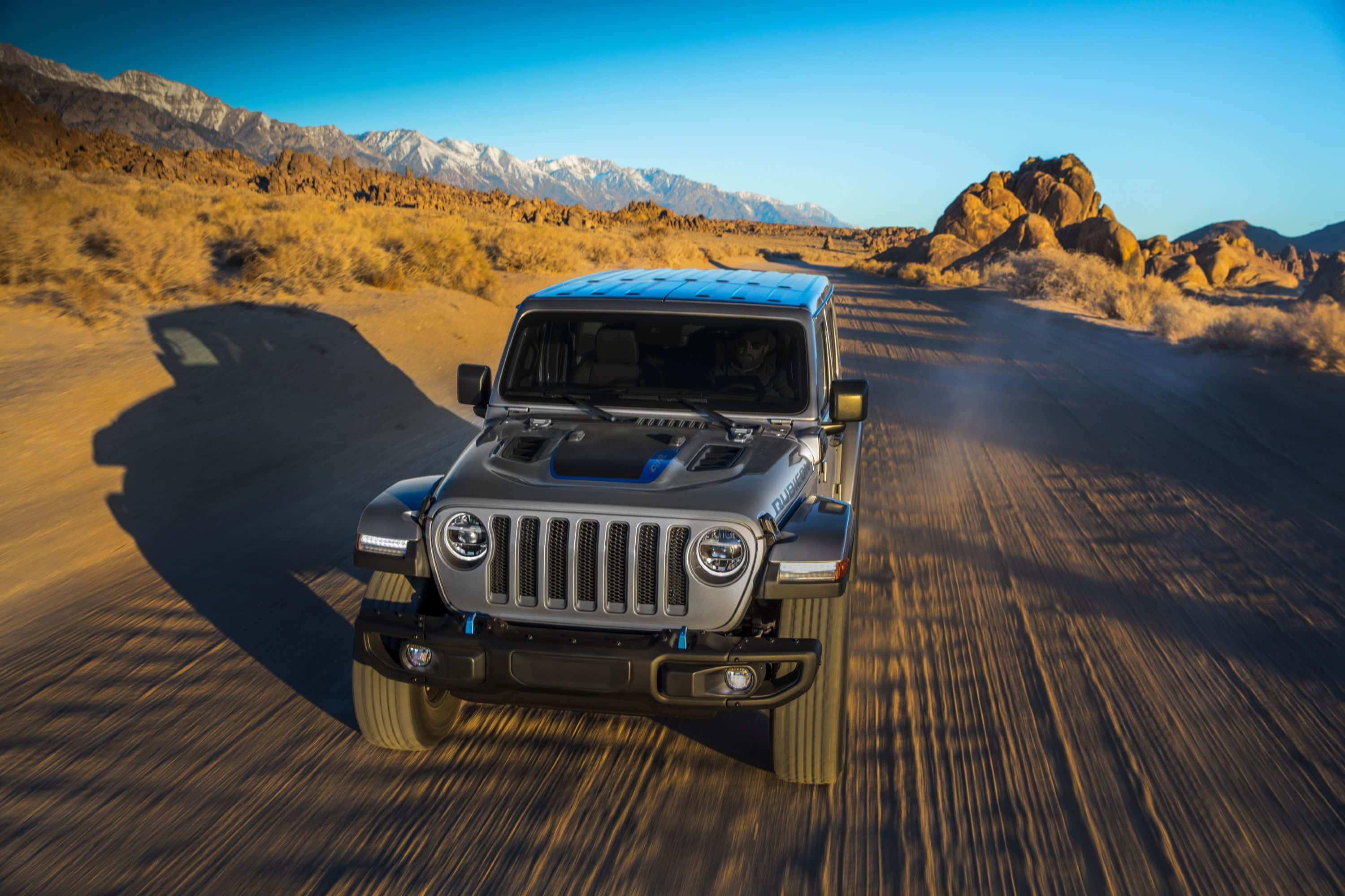 Jeep 動圖 桌布 壁紙 — 一輛銀色Jeep Wrangler在沙漠公路上飛馳，背景是白雪覆蓋的山脈，天空清澈湛藍。
