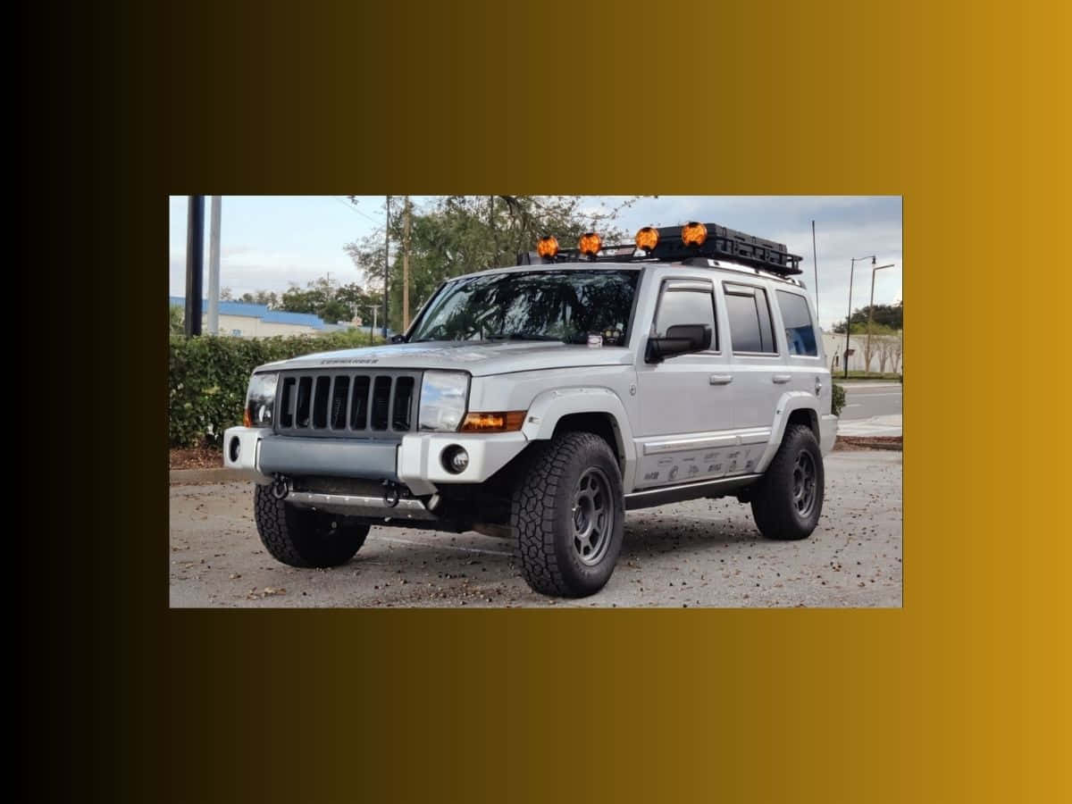 Jeep Commander 桌布 壁紙 — 一輛銀色Jeep Wrangler，帶有橙色車頂燈和堅固輪胎，停泊在碎石上，靠近綠色灌木叢和一座建築物。