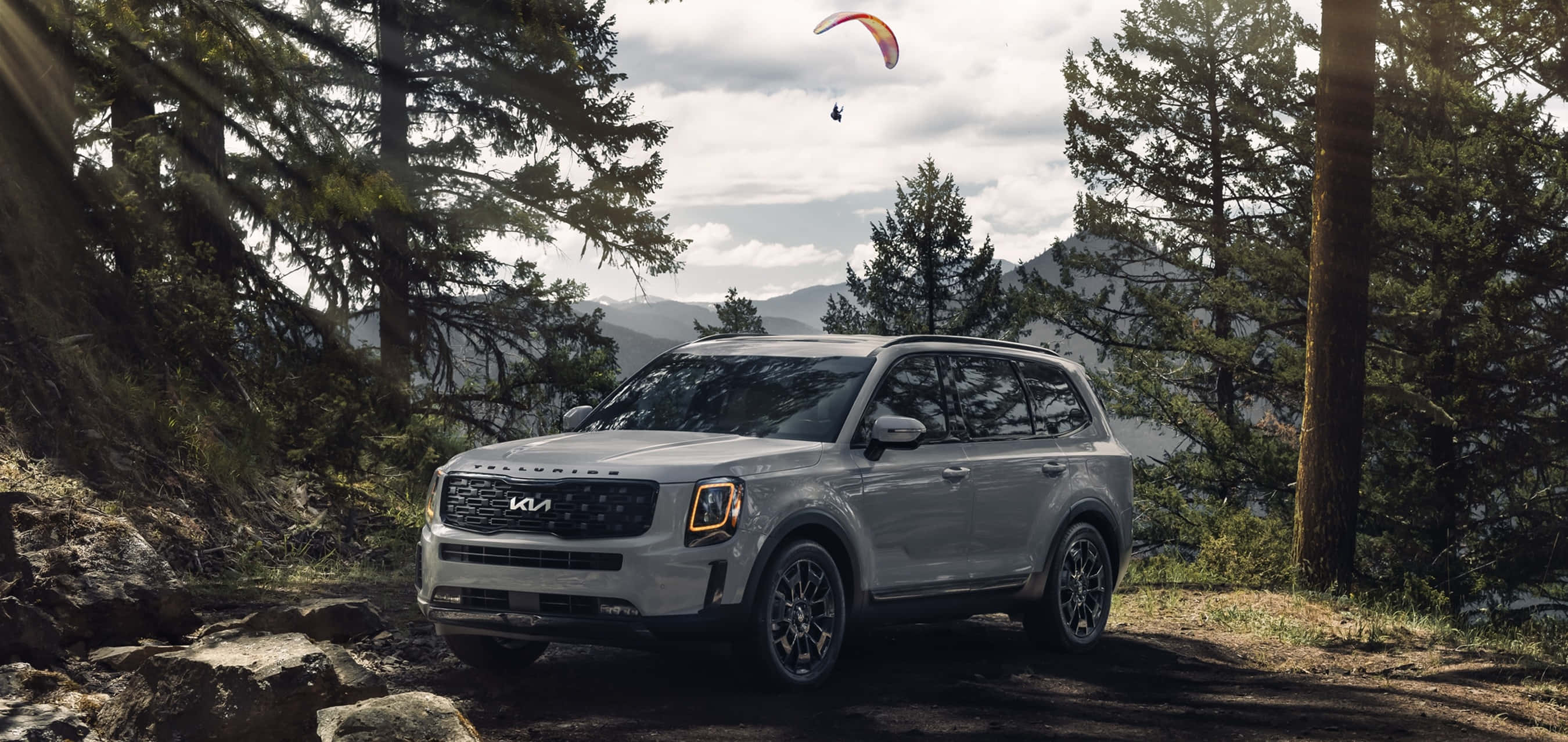 Kia Telluride 桌布 壁紙 — 一輛銀色Kia SUV停泊在森林小徑上，滑翔傘在松樹和遠山上方翱翔。