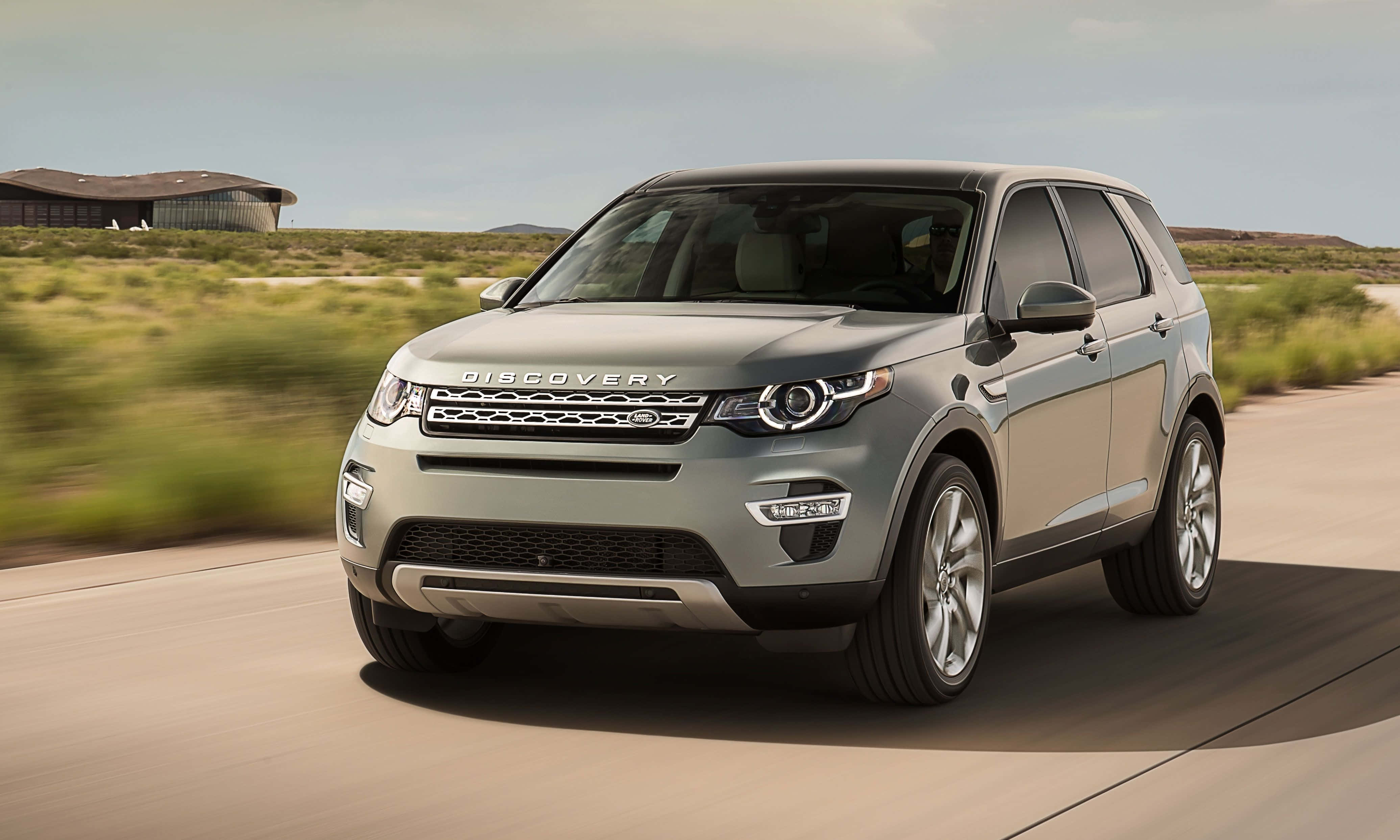 Land Rover Discovery 桌布 壁紙 — 一輛銀色Land Rover Discovery SUV在沙漠公路上疾馳，背景有模糊的綠植和現代建築。