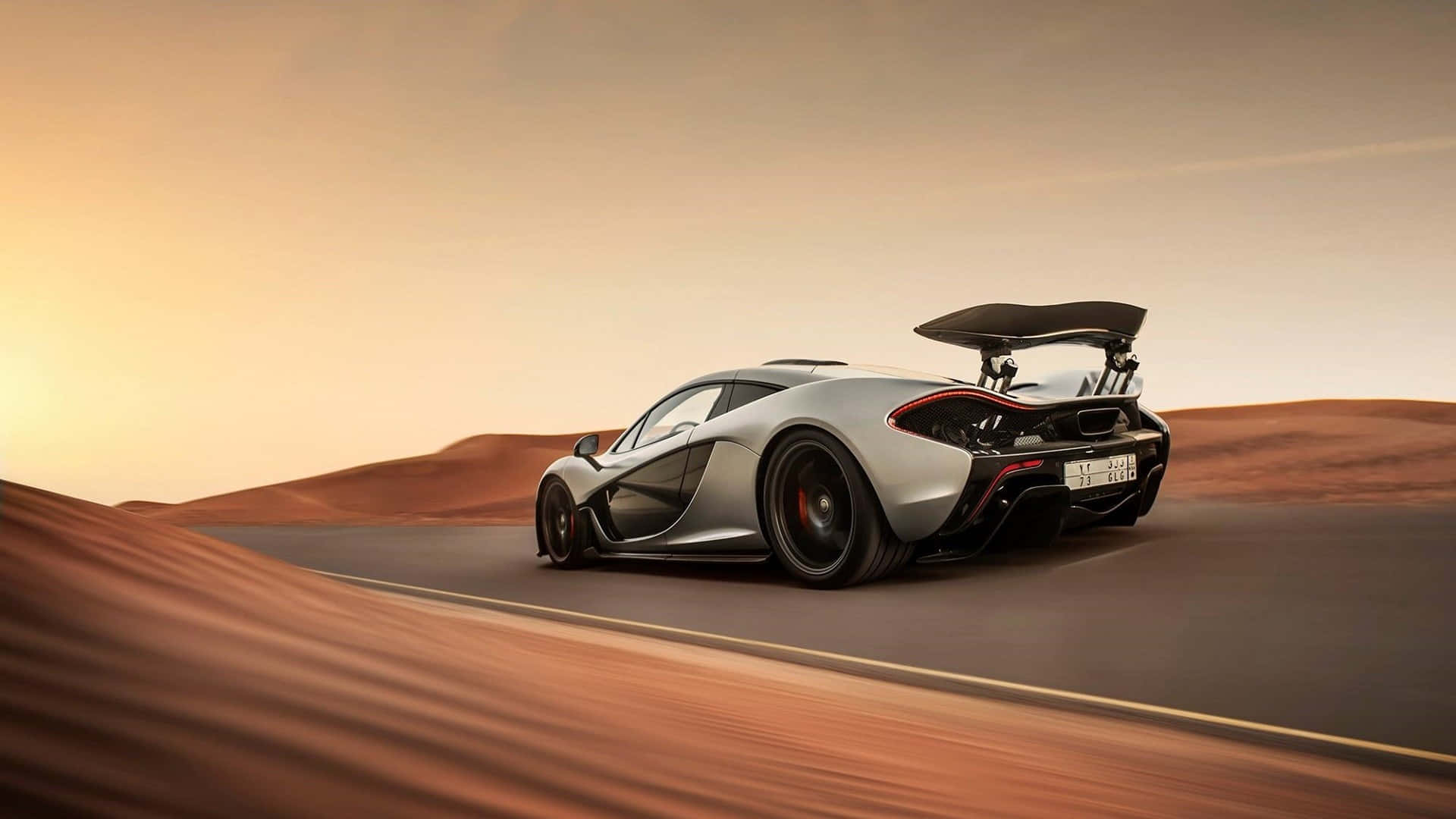 麥拿侖 桌布 壁紙 — 一輛銀色McLaren P1超級跑車在日落時分沿沙漠公路飛馳，帶有大型後擾流板和模糊的背景。