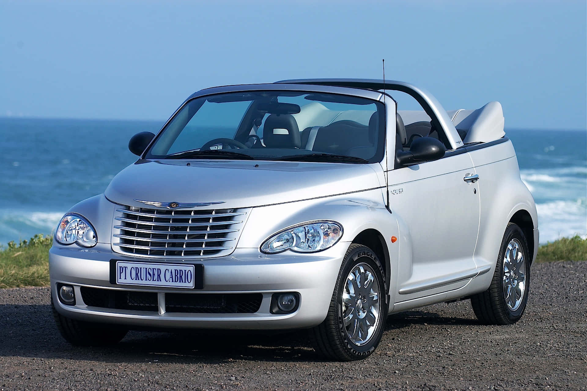 Chrysler Pt Cruiser 桌布 壁紙 — 一輛銀色PT Cruiser Cabrio開篷車停泊在海岸碎石路上，背後是清澈藍天下拍岸的海浪。