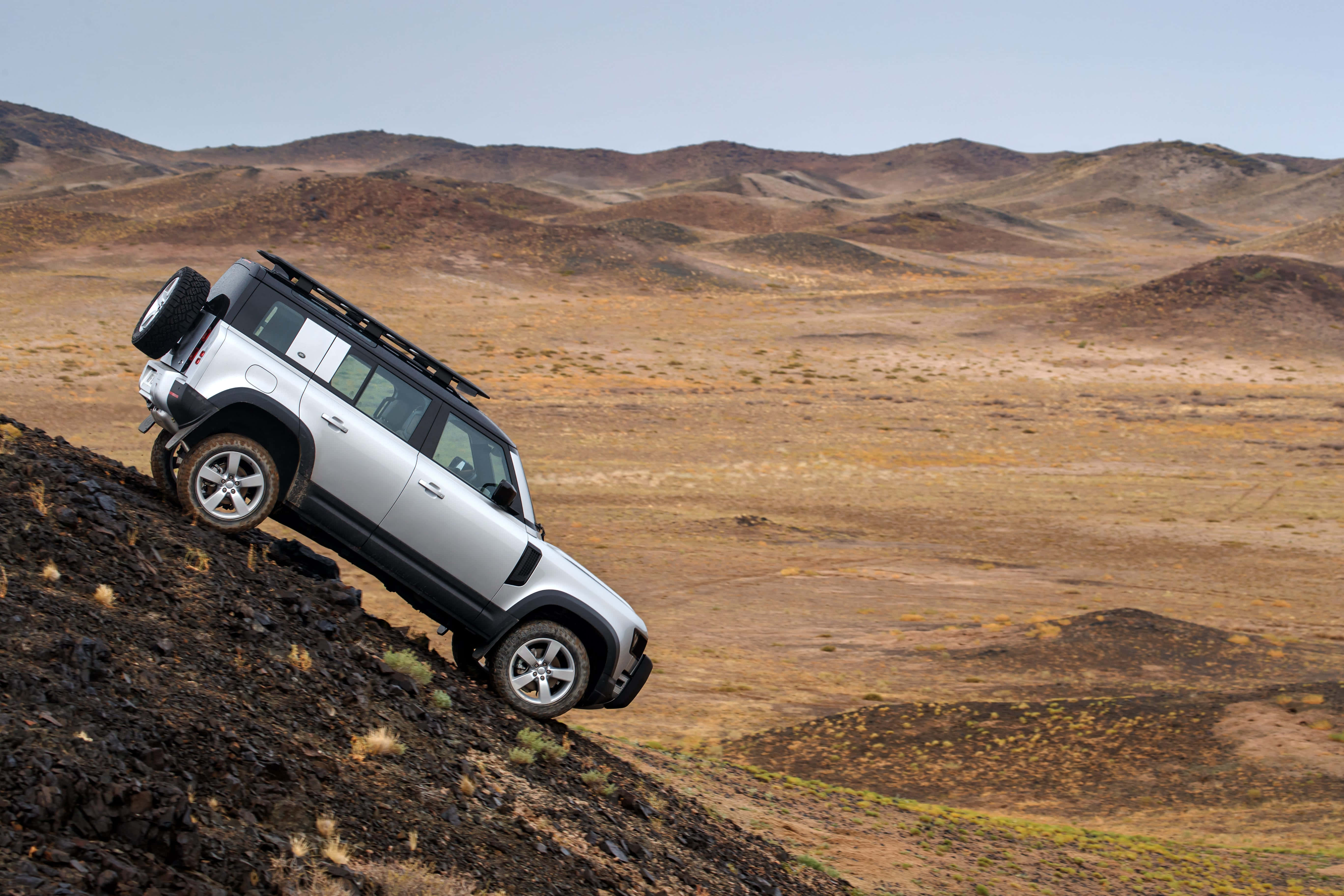 Land Rover Defender 桌布 壁紙 — 一輛銀色SUV在晴朗天空下征服廣闊乾旱沙漠景觀中的陡峭岩石斜坡。