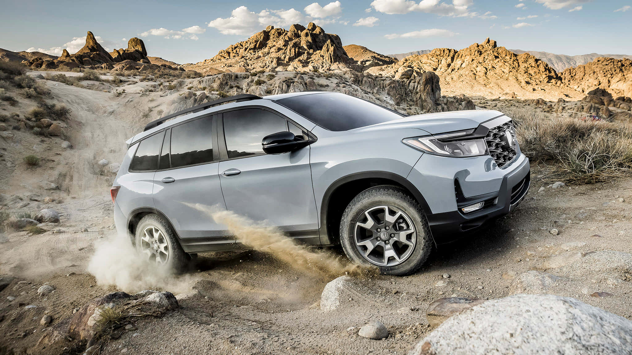 Honda Passport 桌布 壁紙 — 一輛銀色SUV在部分多雲的天空下，駛過岩石沙漠地形，背景是鋸齒狀的山脈，揚起灰塵。