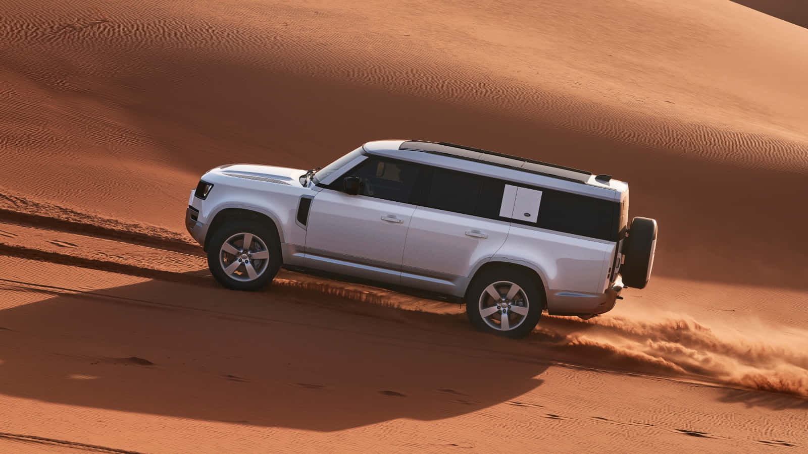 Land Rover Defender 桌布 壁紙 — 一輛銀色SUV在金色沙漠沙丘中疾馳，揚起沙塵，沐浴在陽光下。