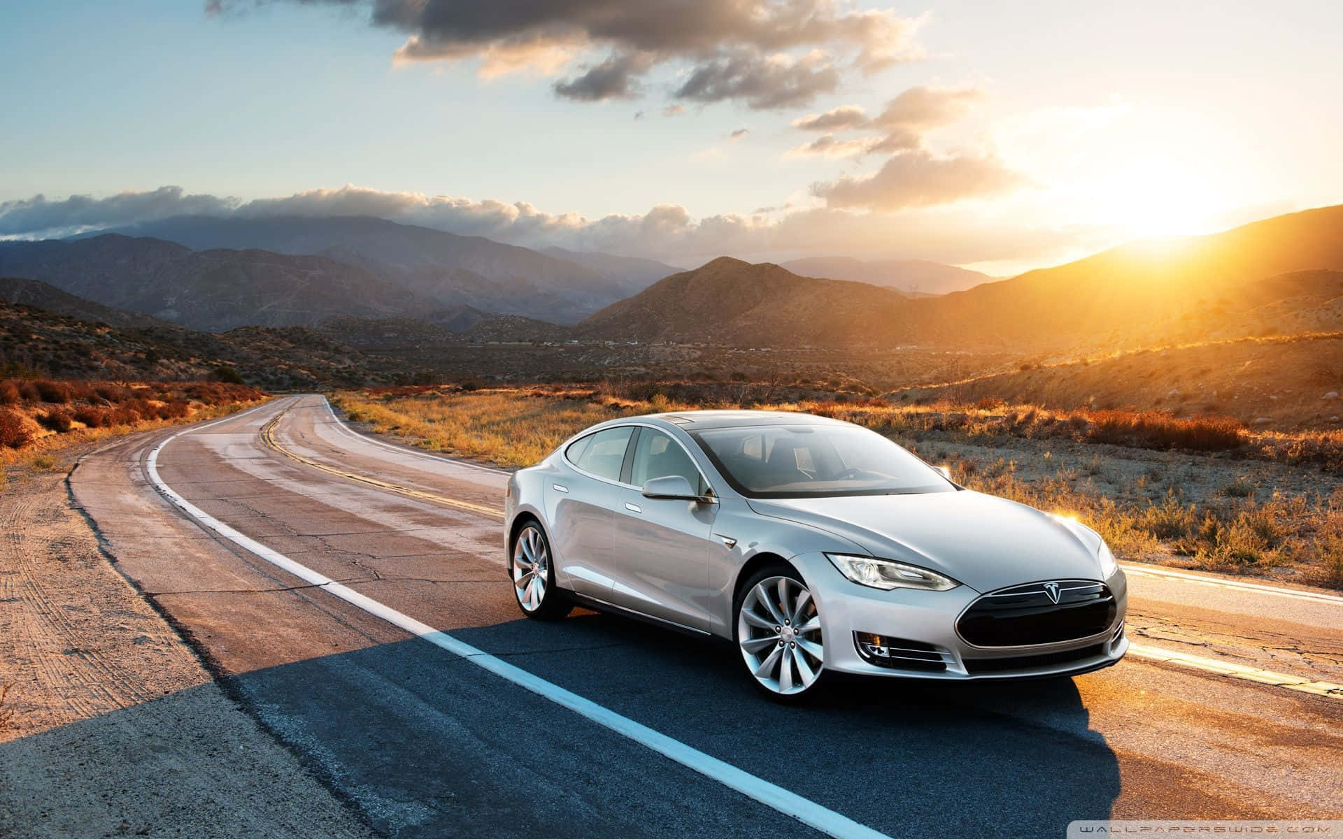 Hybrid Car 桌布 壁紙 — 一輛銀色Tesla Model S沿著蜿蜒的山路行駛，沐浴在金色的夕陽光芒中。