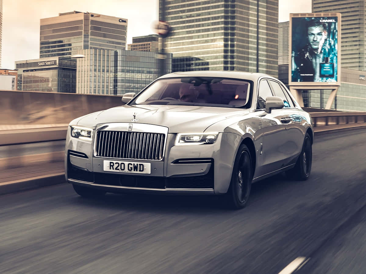 Rolls Royce 桌布 壁紙 — 一輛銀色勞斯萊斯 Ghost 在都市高架橋上奔馳，背景是玻璃摩天大樓和 Chanel 廣告牌，呈現模糊動態效果。