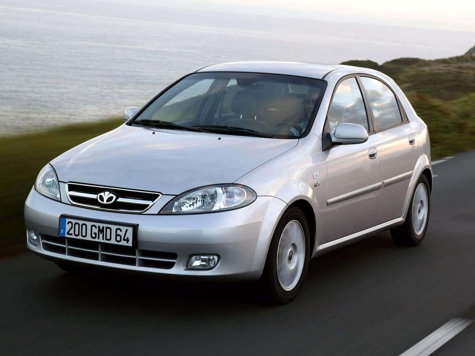 Daewoo 桌布 壁紙 — 一輛銀色大宇Matiz沿著海岸道路行駛，背景是模糊的綠色山丘和海洋。