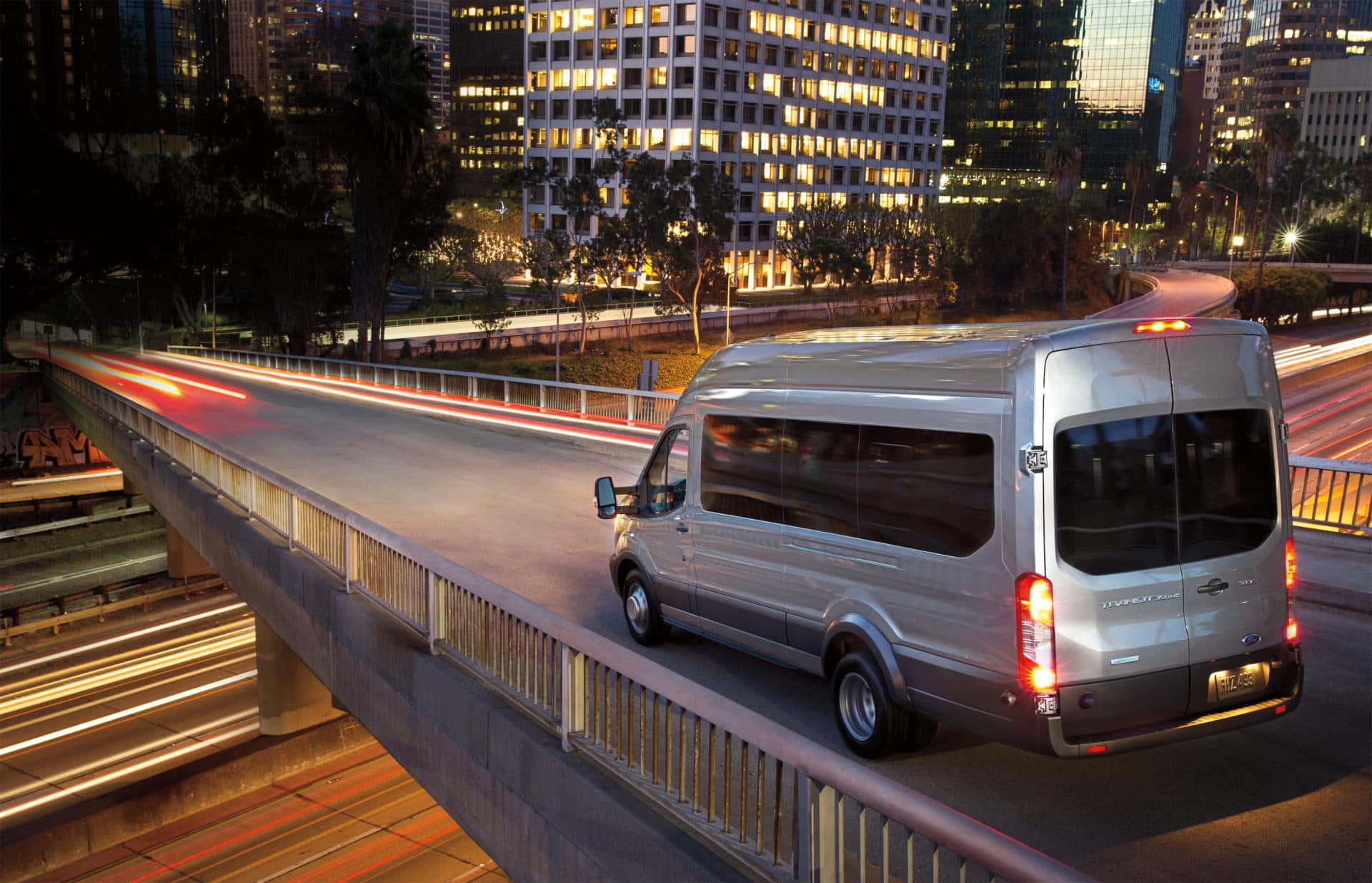 Ford Transit 桌布 壁紙 — 一輛銀色客貨車在夜間行駛於高架高速公路，背景是流動的城市燈光和燈火通明的摩天大樓。
