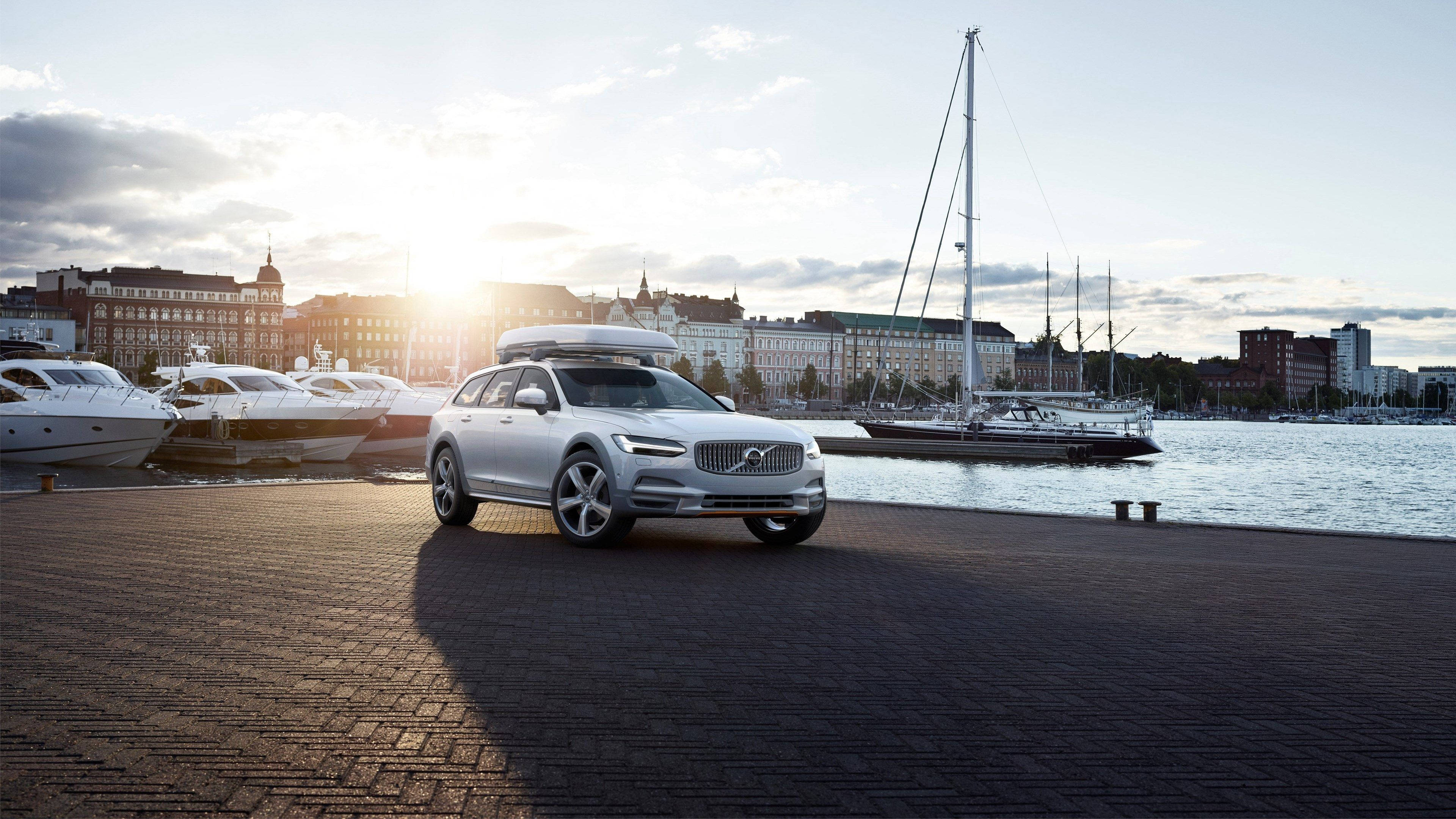 Volvo V90 Cross Country 壁紙 — 一輛銀色富豪旅行車連車頂箱停泊在磚碼頭上，夕陽西下，旁邊有帆船和城市建築。