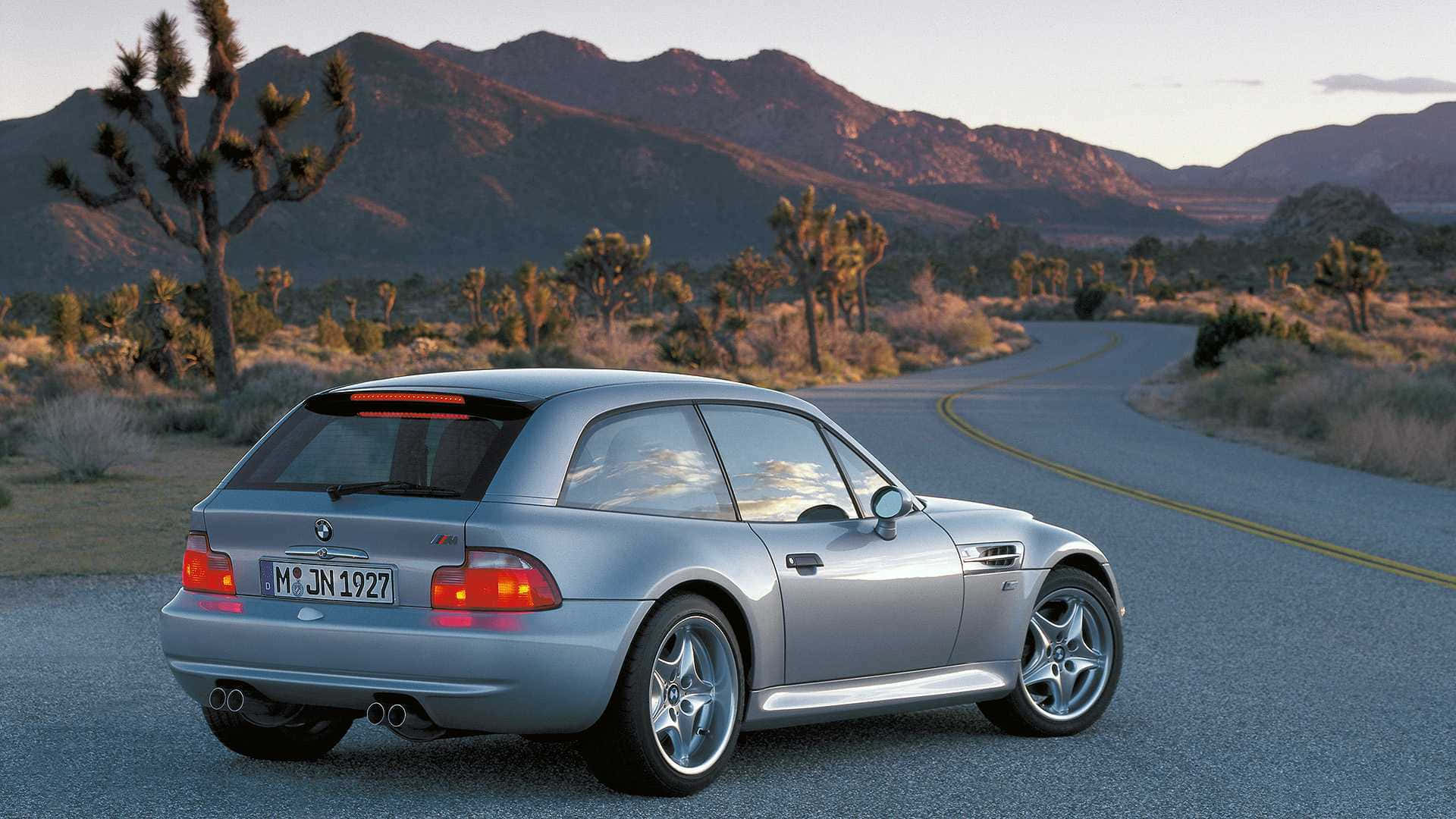 Bmw Z3 桌布 壁紙 — 一輛銀色寶馬M3雙門跑車在蜿蜒的沙漠公路上行駛，兩旁有約書亞樹和山脈，天空呈現柔和的日落色彩。