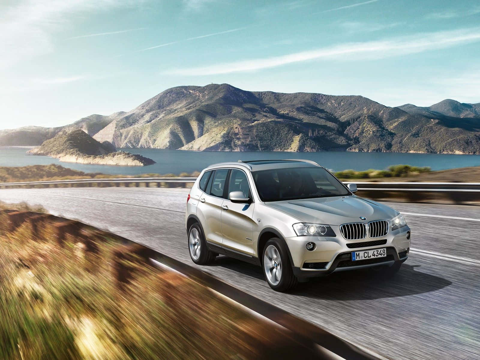 Bmw X3 桌布 壁紙 — 一輛銀色寶馬SUV在晴朗藍天下沿著蜿蜒的海岸道路行駛，背景是山脈和湖泊。