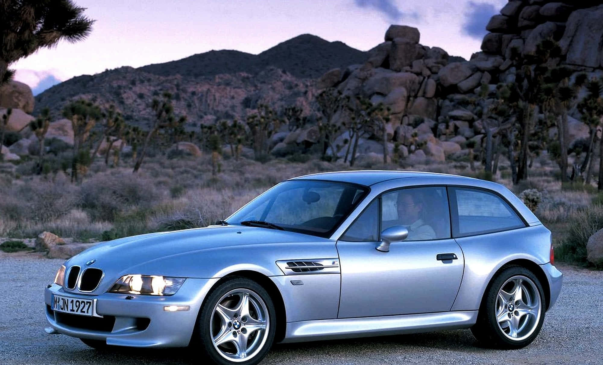 Bmw Z3 桌布 壁紙 — 一輛銀色寶馬Z3開篷跑車在暮色天空下閃閃發光，停泊在約書亞樹遍佈的岩石沙漠地帶的碎石路上。