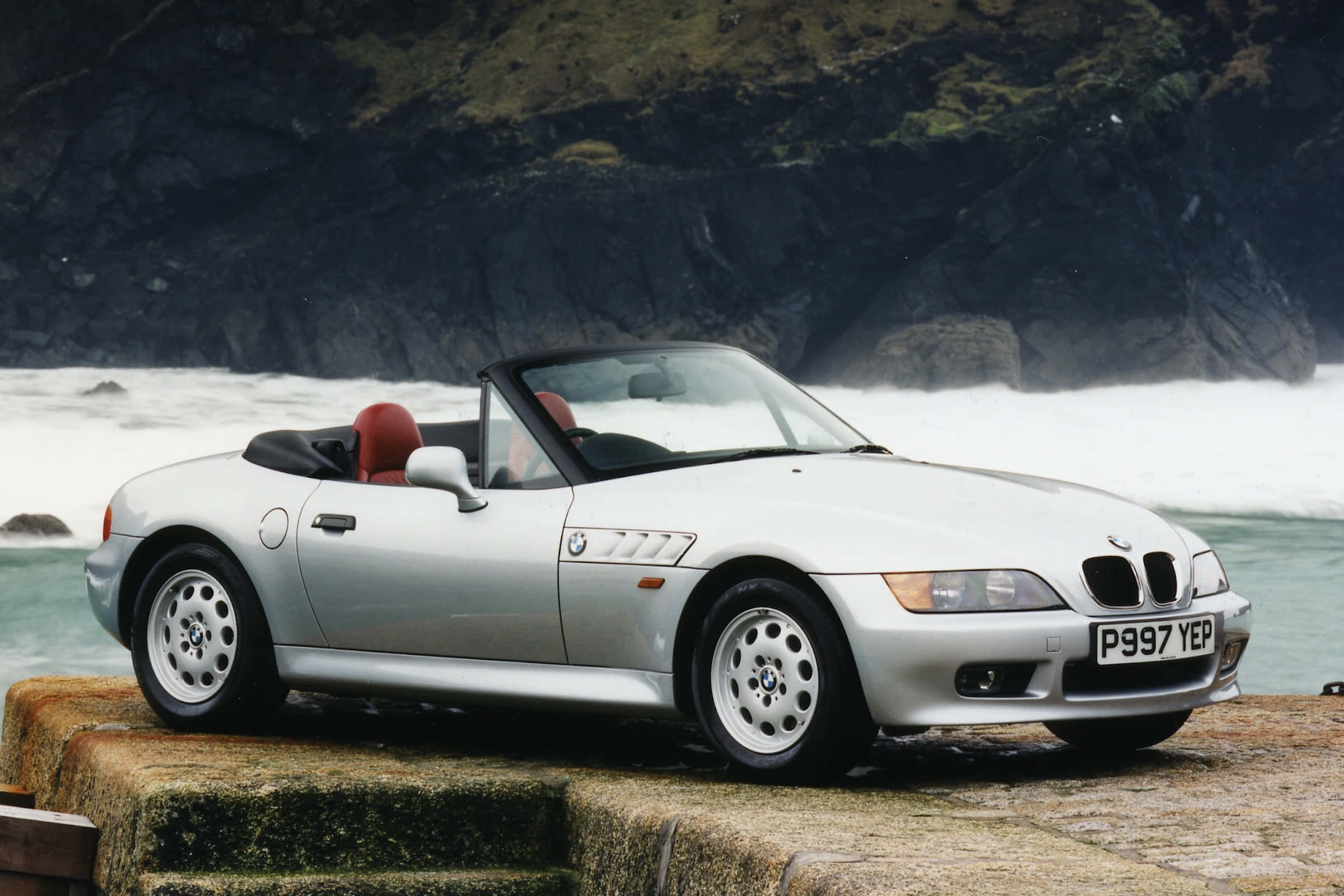 Bmw Z3 桌布 壁紙 — 一輛銀色寶馬Z3開篷跑車配紅色座椅，停泊在石上，旁邊是拍打暗黑懸崖的洶湧海浪。