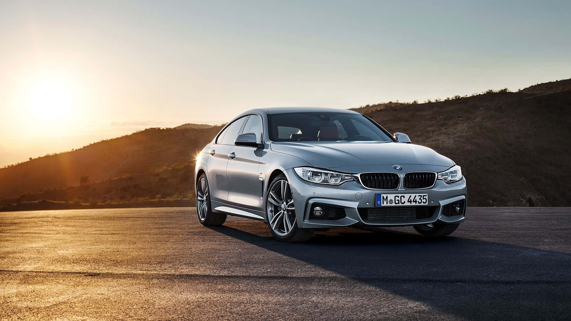 Bmw 4 Series 桌布 壁紙 — 一輛銀色寶馬房車在金色夕陽光下，於沙漠公路上閃耀，背景是起伏的山丘。
