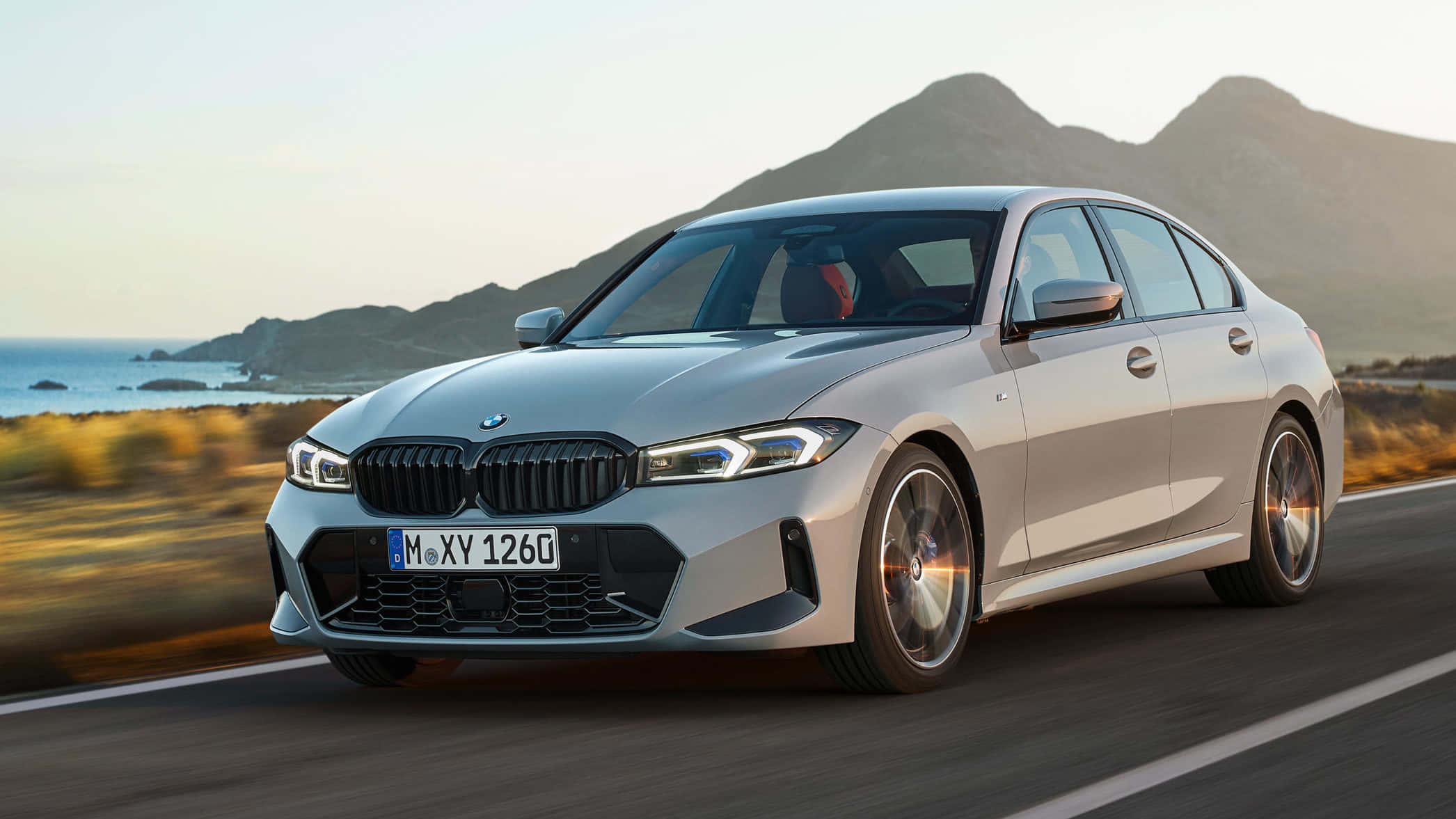 Bmw 3 Series 桌布 壁紙 — 一輛銀色寶馬房車在黃金時段沿著海岸公路疾馳，背景是山脈和海洋。