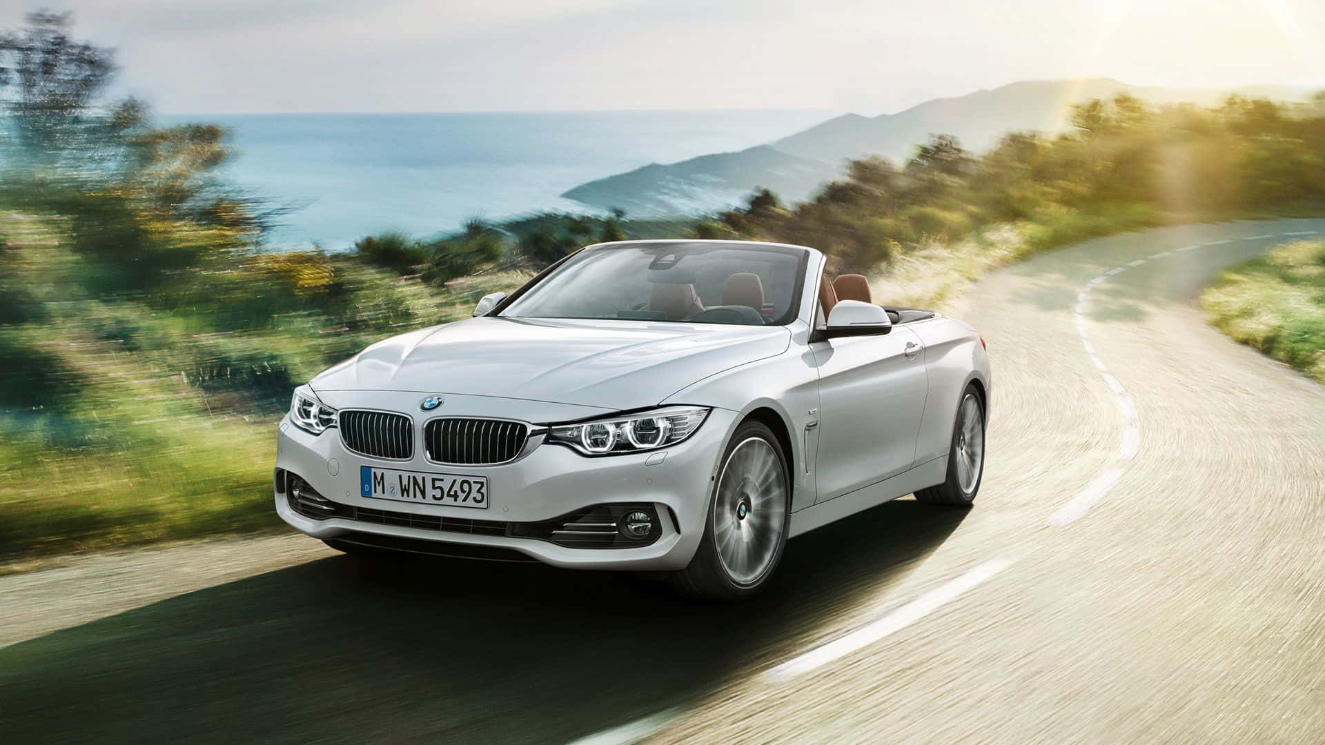 Bmw 4 Series 桌布 壁紙 — 一輛銀色寶馬開篷跑車在明亮陽光下，沿著海岸公路飛馳，背景是模糊的綠化和海景。