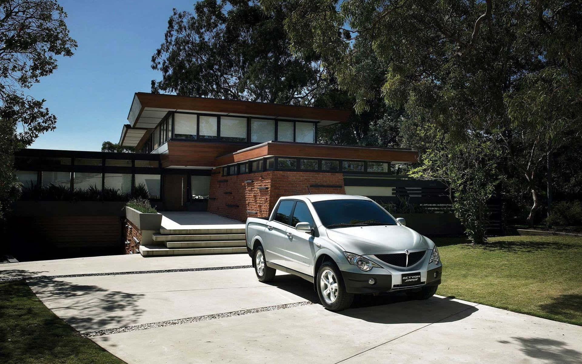 Ssangyong 桌布 壁紙 — 一輛銀色小型貨車停泊在現代木磚房屋前的混凝土車道上，周圍綠樹成蔭，天空湛藍。