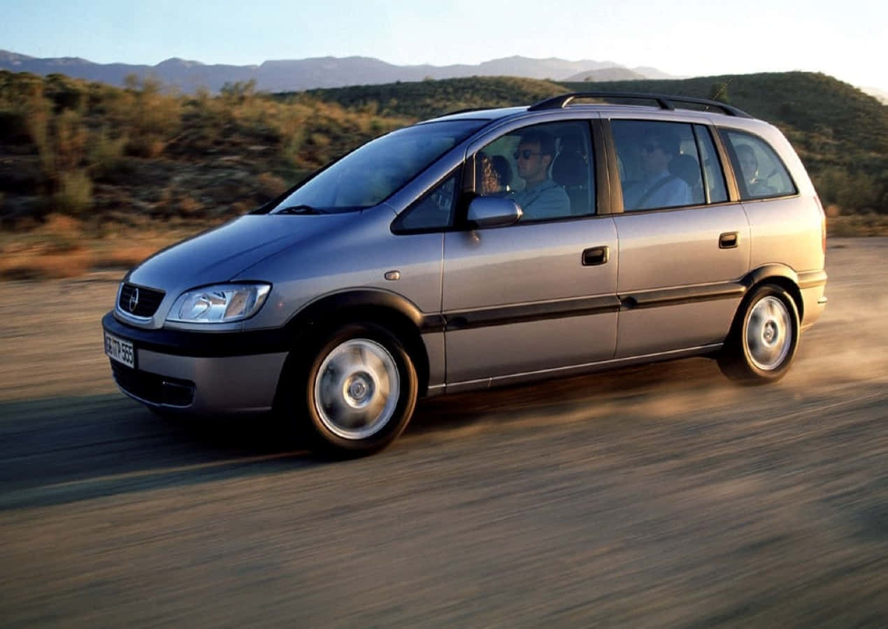 Opel Zafira 桌布 壁紙 — 一輛銀色小型貨車在晴朗天空下，以山脈為背景，在塵土飛揚的道路上疾馳。