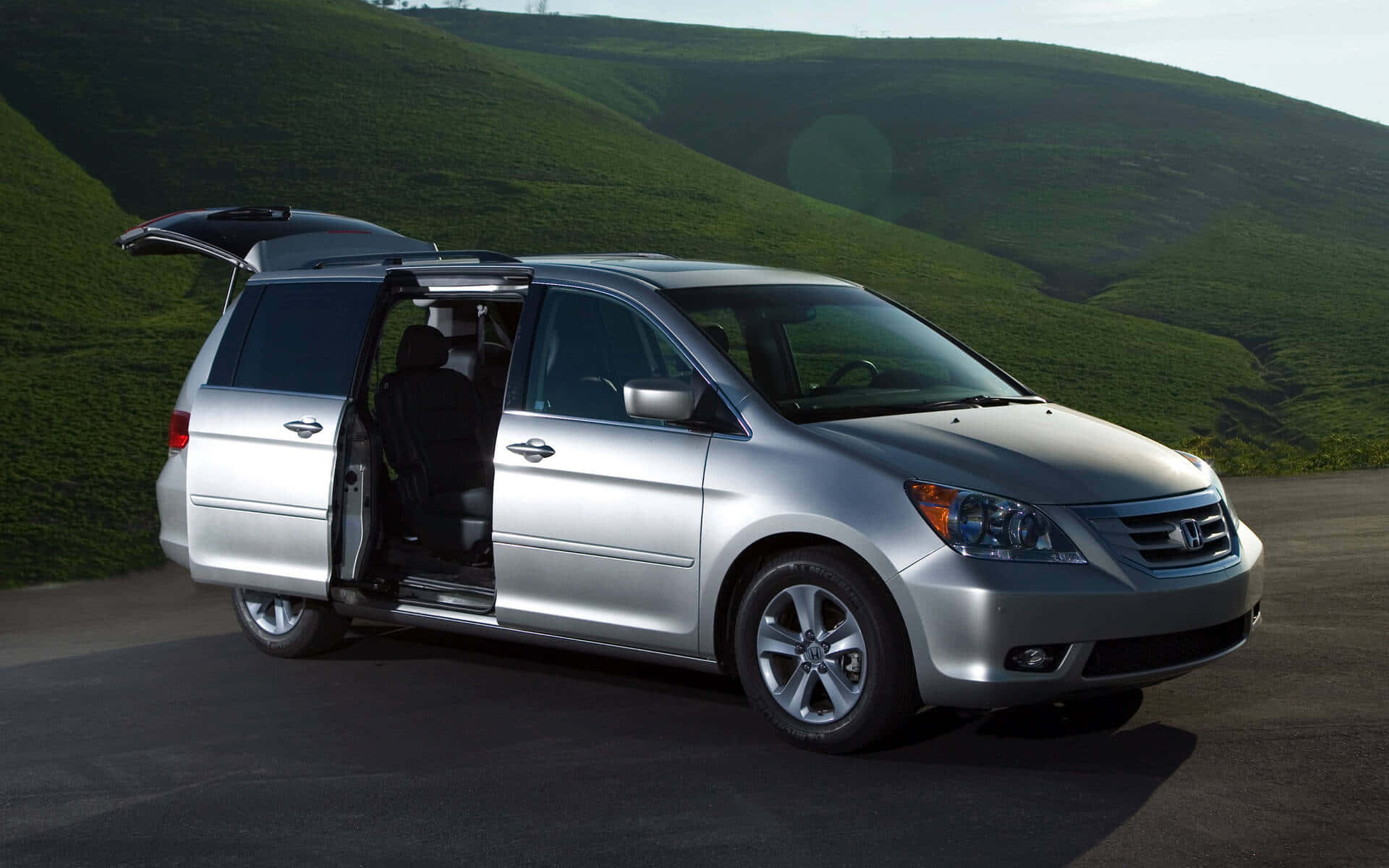 Honda Odyssey 桌布 壁紙 — 一輛銀色小型貨車打開滑門，停在鋪砌的道路上，背景是起伏的綠色山丘，光線明亮。