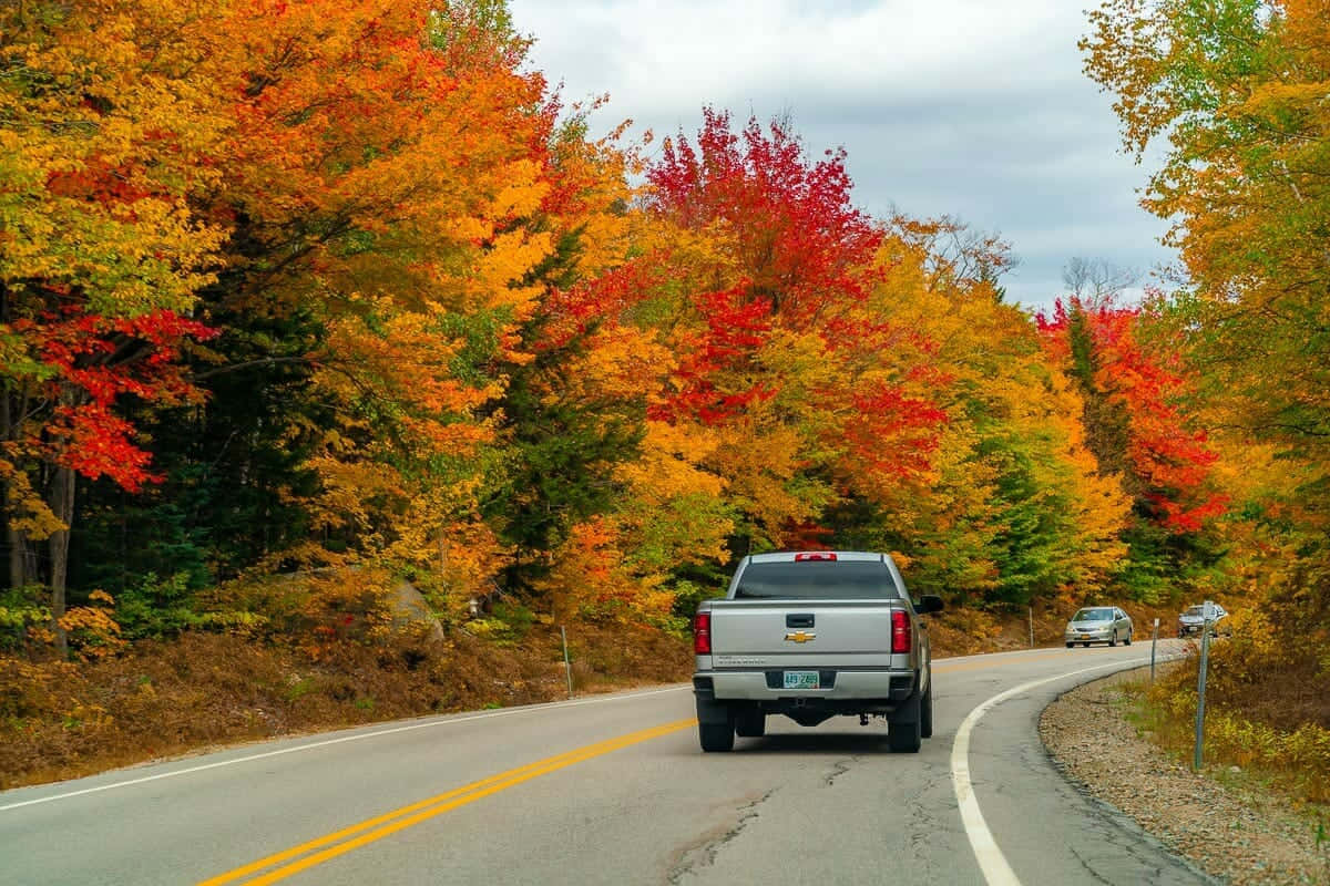 Fall Road 桌布 壁紙 — 一輛銀色小型貨車沿著蜿蜒的道路行駛，道路兩旁是色彩繽紛的秋樹，呈現火紅、橙黃色調，天空多雲。
