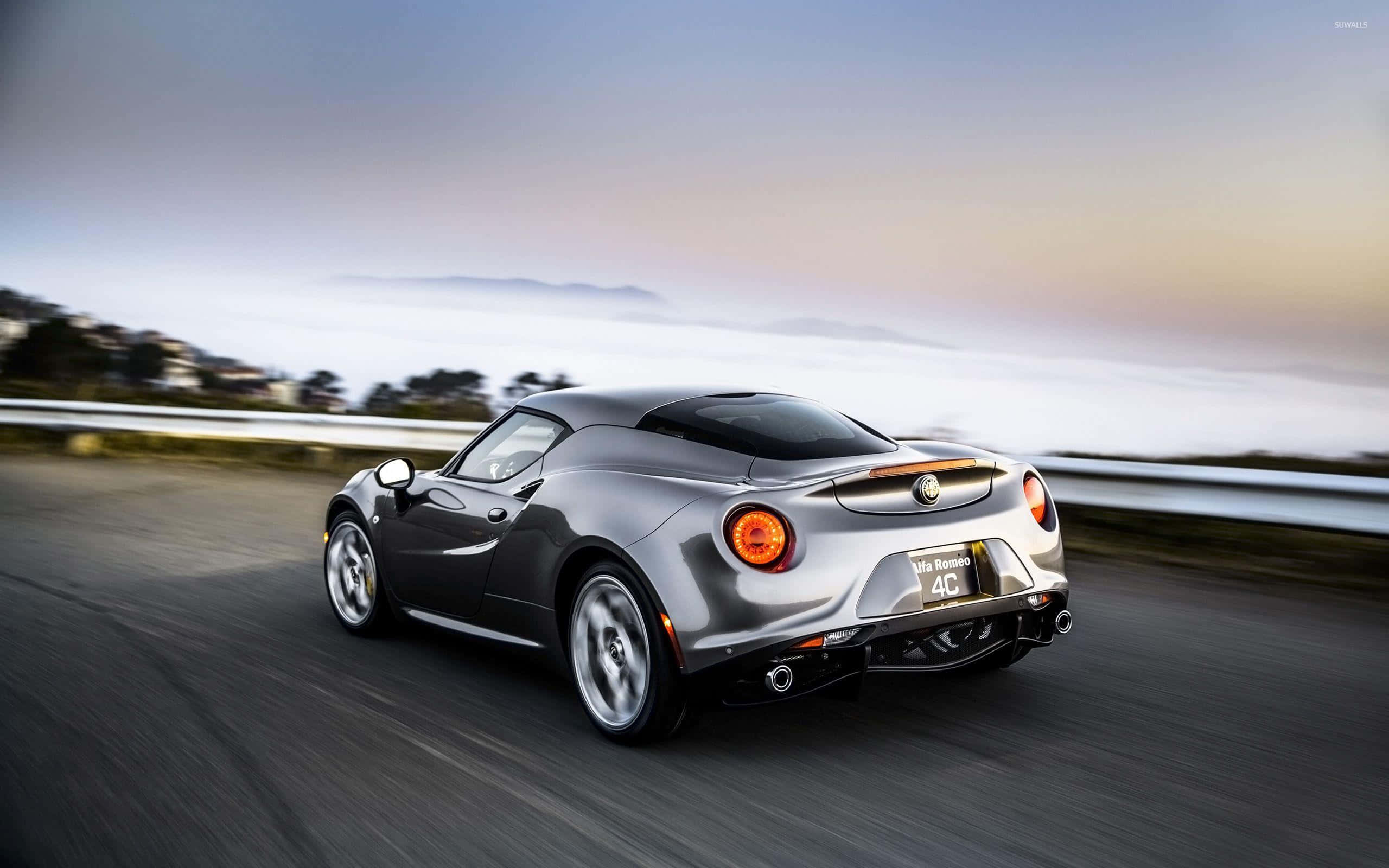 Alfa Romeo 4c 桌布 壁紙 — 一輛銀色愛快羅密歐4C跑車在日落時分沿著海岸公路奔馳，帶有動態模糊和遠山。