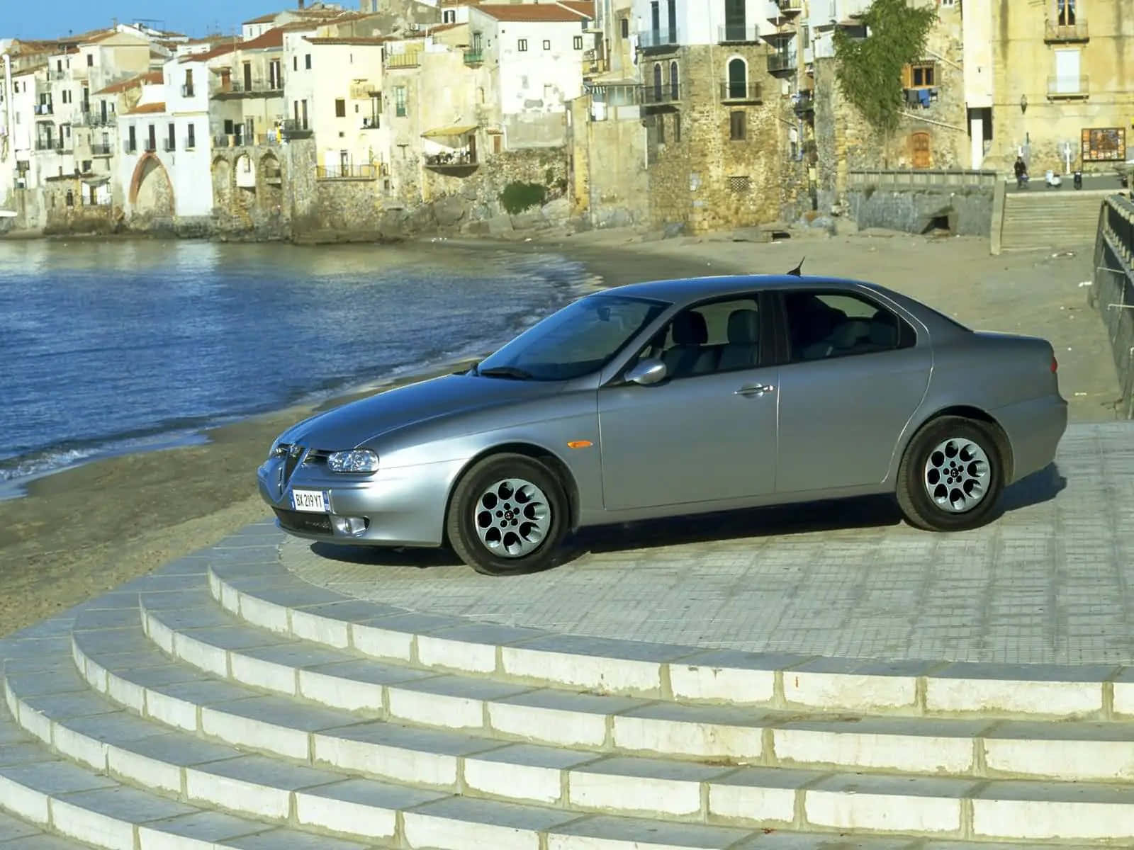 Alfa Romeo 156 桌布 壁紙 — 一輛銀色愛快羅密歐房車停泊在彎曲的石階上，旁邊是陽光普照的海邊小鎮，有陶土屋頂和寧靜的藍色海水。