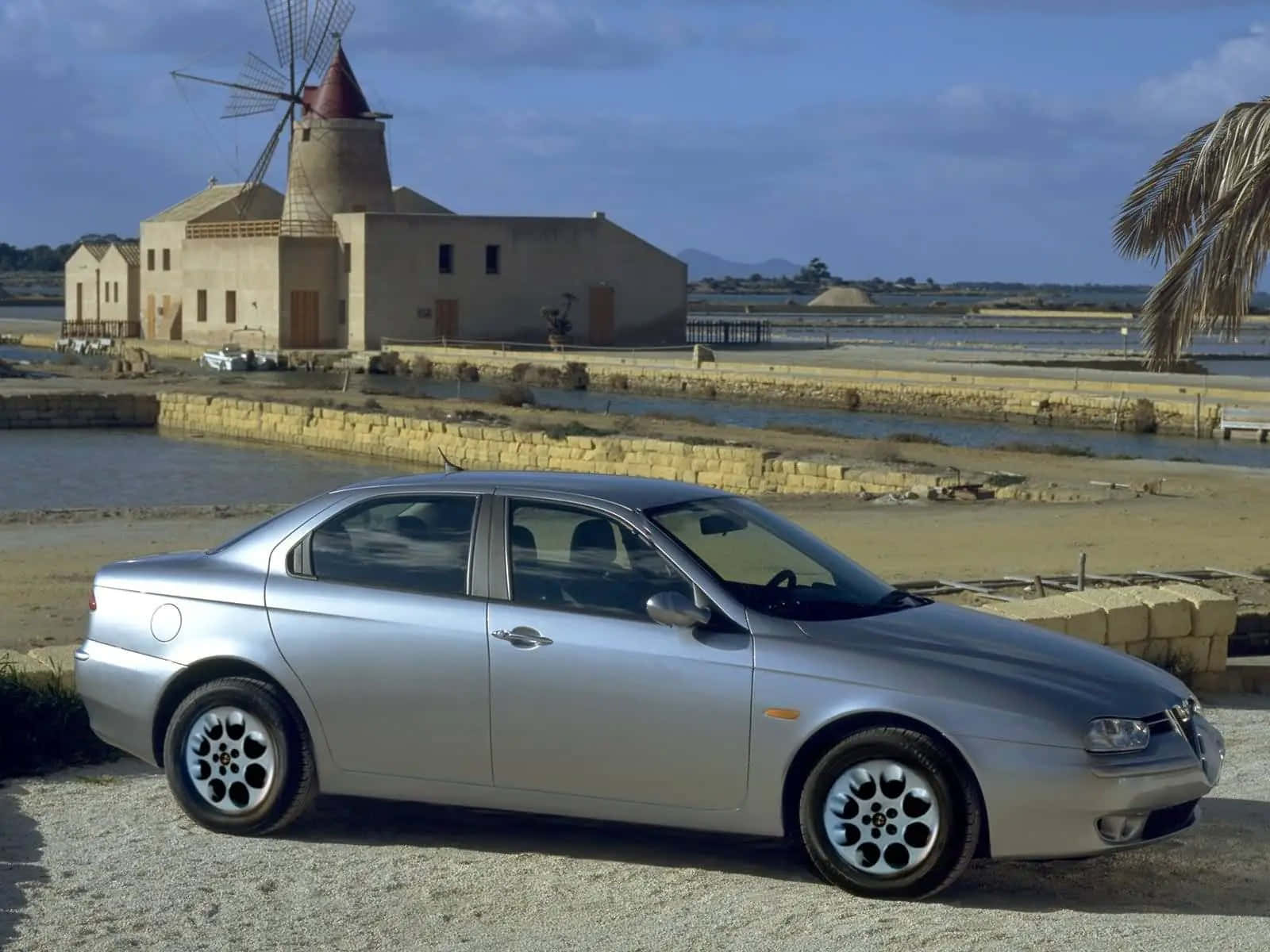 Alfa Romeo 156 桌布 壁紙 — 一輛銀色愛快羅密歐房車停泊在碎石路上，靠近歷史風車和鹽田，天空湛藍。