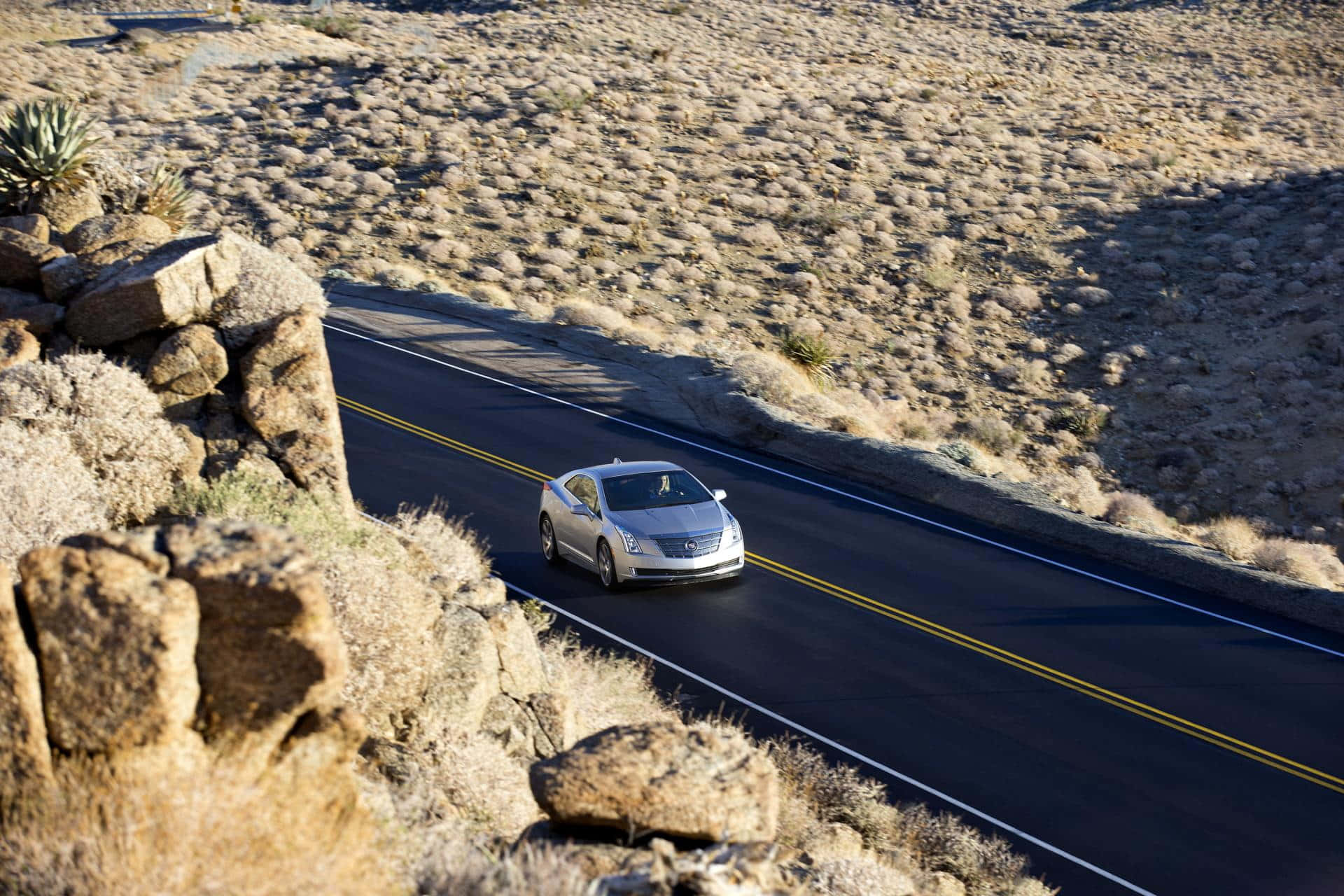 Cadillac Elr 桌布 壁紙 — 一輛銀色房車在明亮的日光下行駛在一條蜿蜒的沙漠公路上，兩旁是岩石地形和乾燥的灌木叢。