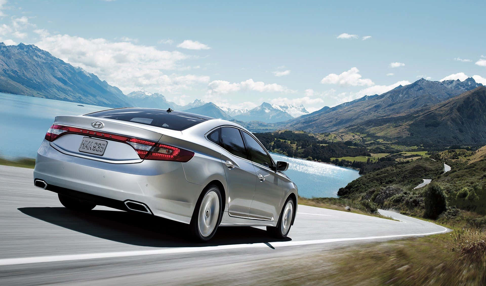 Hyundai Azera 桌布 壁紙 — 一輛銀色房車在晴朗的天空下，沿著蜿蜒的山路行駛，旁邊是寧靜的藍色湖泊。