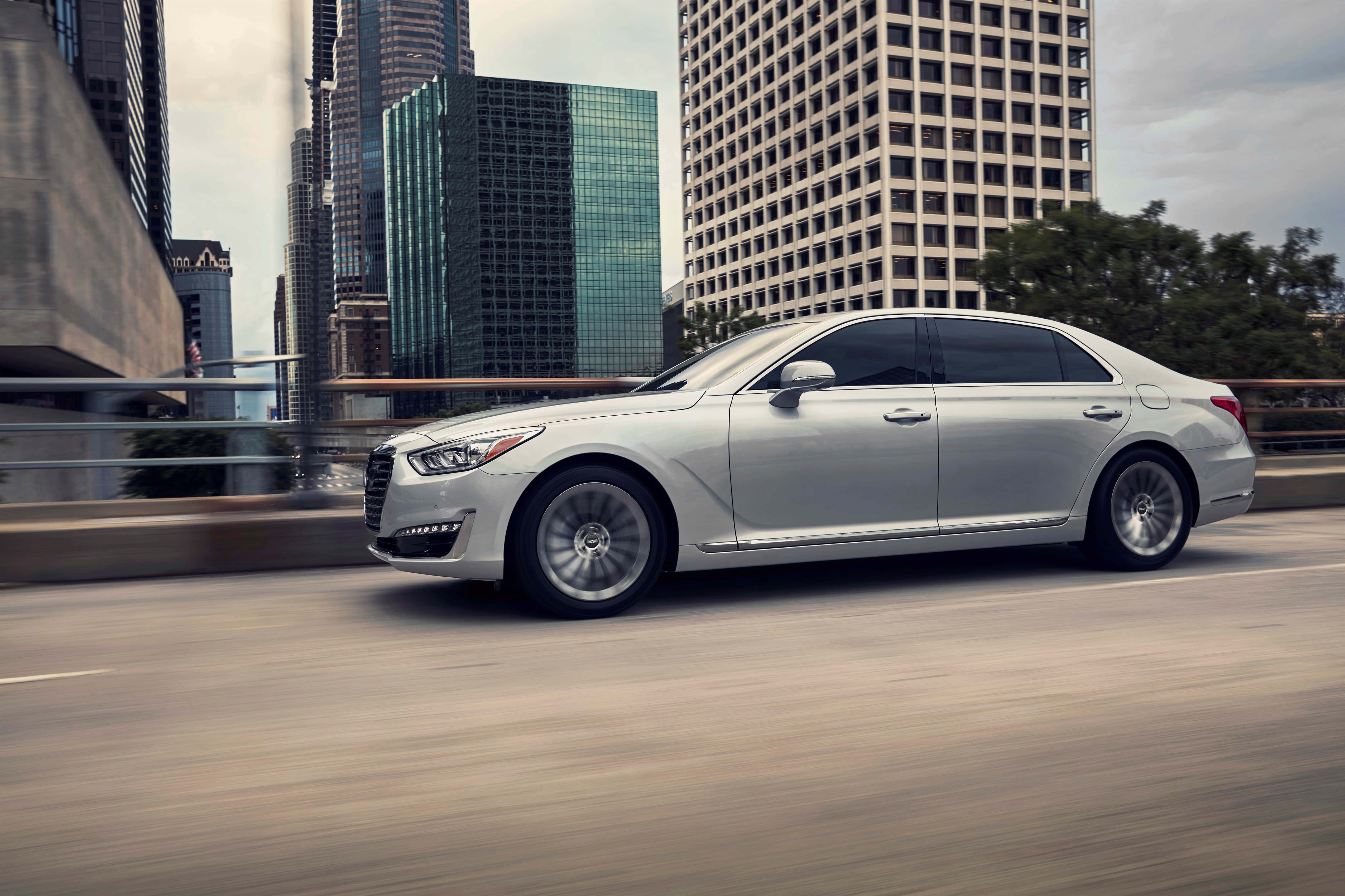 Genesis G90 桌布 壁紙 — 一輛銀色房車在都市橋樑上飛馳，背景是高樓大廈和綠化。
