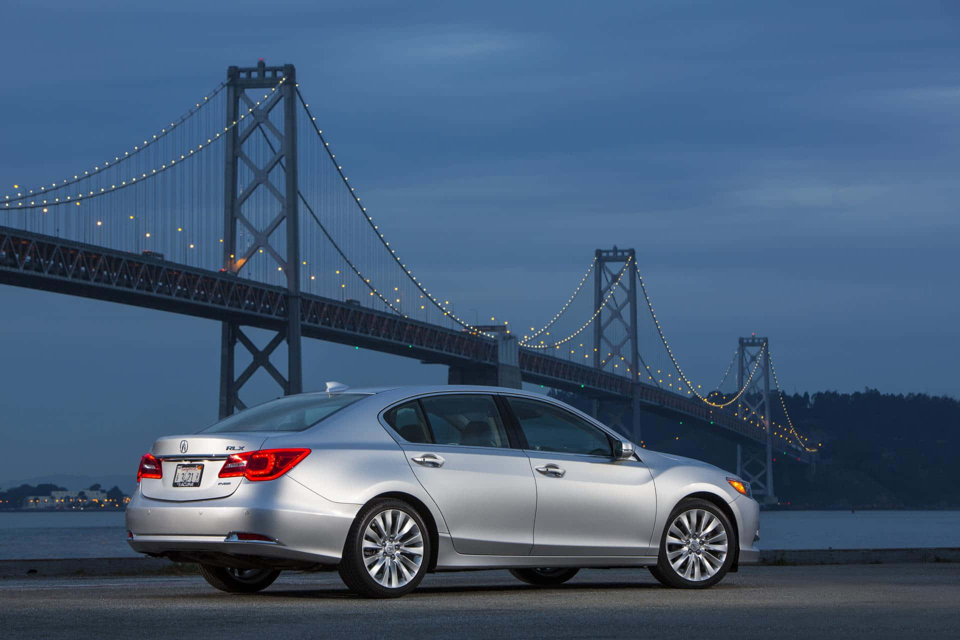 Acura Rlx 桌布 壁紙 — 一輛銀色房車在黃昏時停泊，身後是燈火通明的海灣大橋，矗立於平靜的水面上。