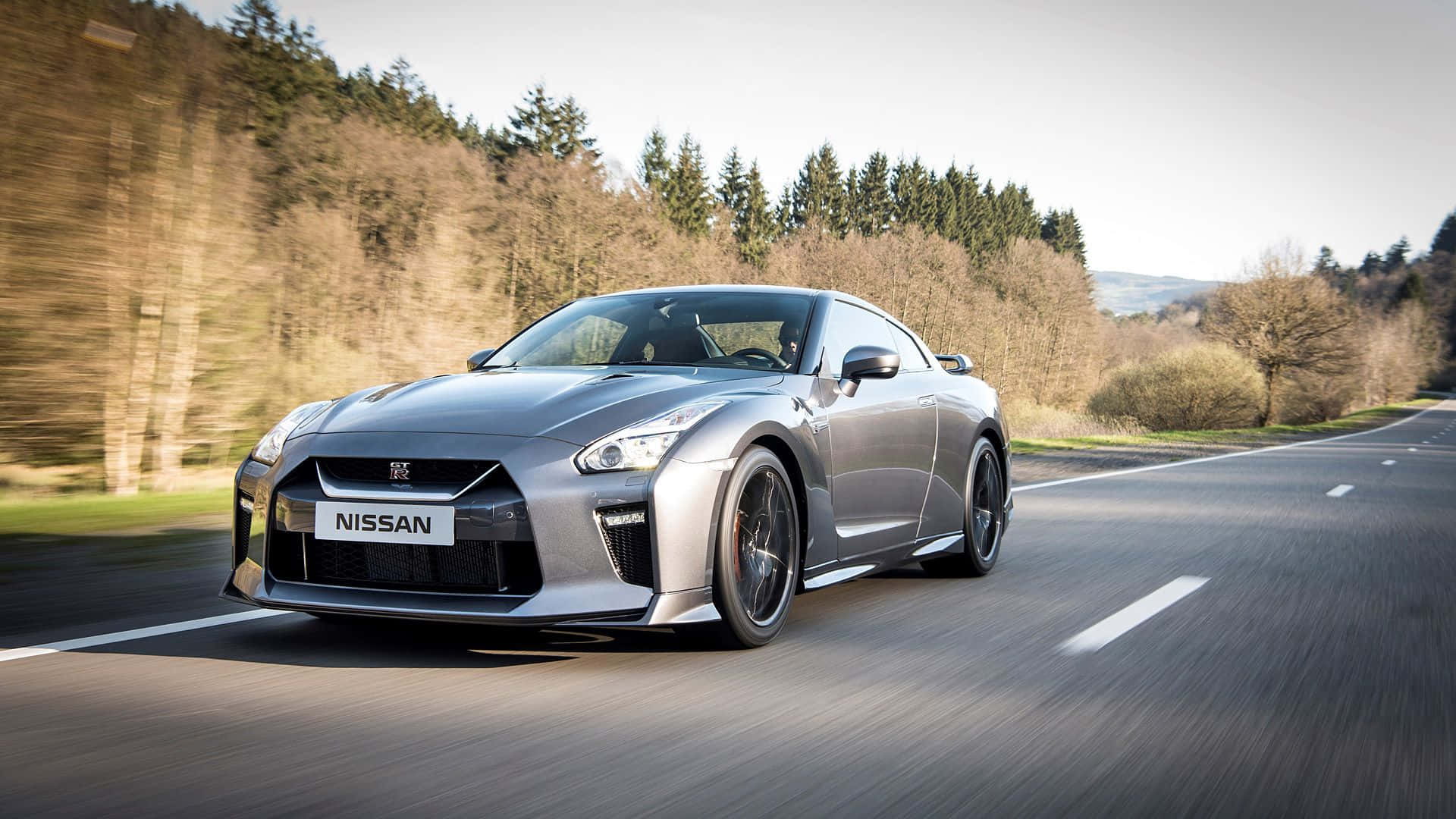 Gtr 桌布 壁紙 — 一輛銀色日產GT-R在蜿蜒的林間道路上疾馳，樹木模糊，天空晴朗。