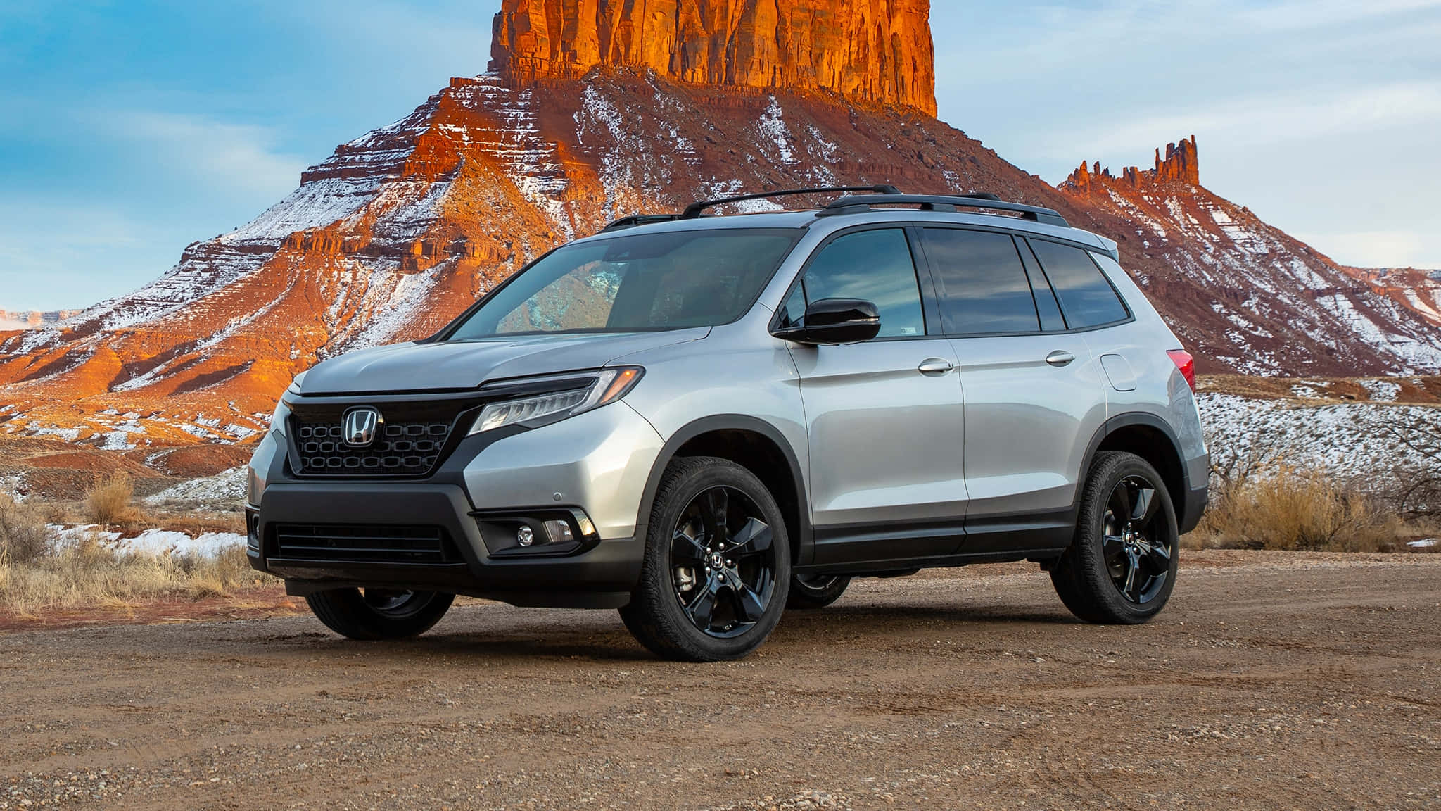 Honda Passport 桌布 壁紙 — 一輛銀色本田Pilot SUV配黑色輪圈，停泊在泥土上，背景是白雪覆蓋的紅色岩層，天空清澈湛藍。