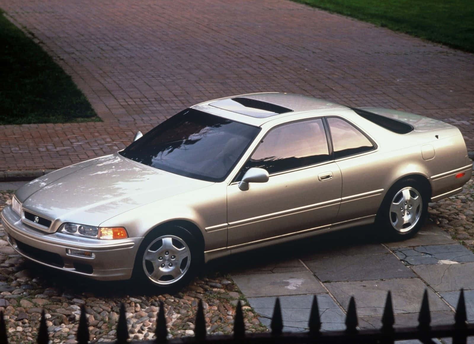 Acura Legend 桌布 壁紙 — 一輛銀色本田轎跑車停泊在鵝卵石車道上，旁邊是磚砌小路，前景有黑色鐵欄杆。