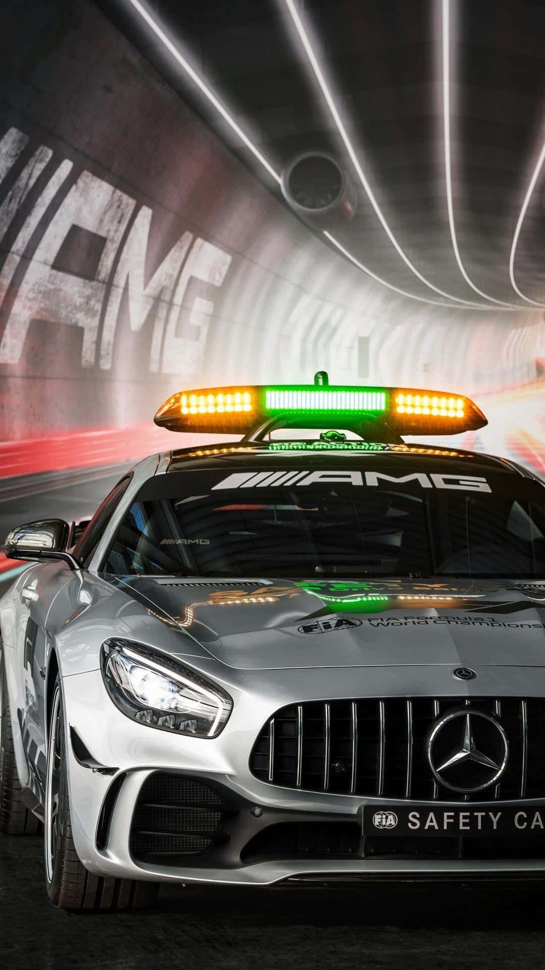 Safety Car 桌布 壁紙 — 一輛銀色梅賽德斯-AMG安全車閃爍著燈光，穿過一條帶有發光霓虹線條和牆上「AMG」塗鴉的隧道。