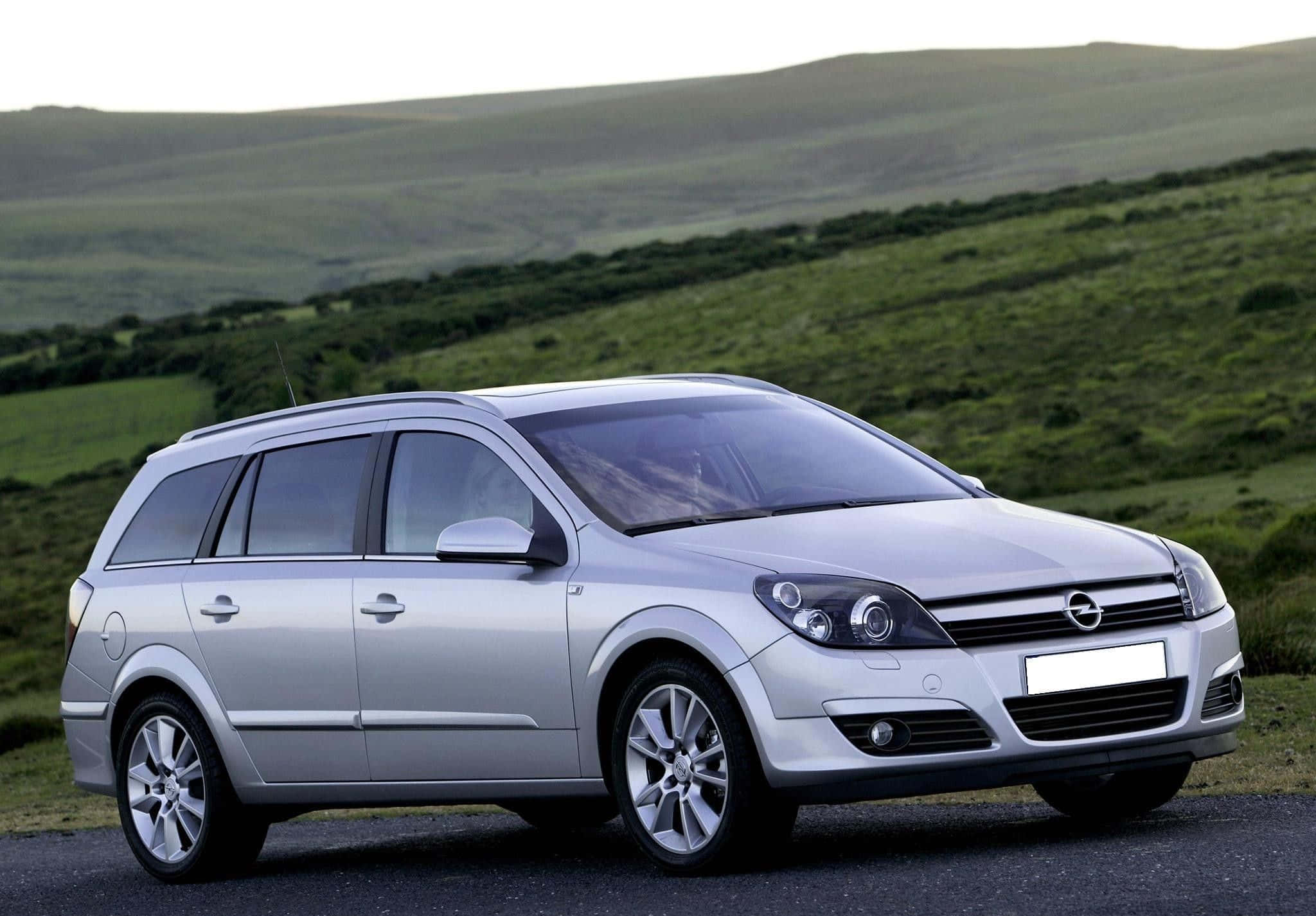 Opel Astra 桌布 壁紙 — 一輛銀色歐寶旅行車在黃昏時停在一條路上，背景是起伏的綠色山丘。