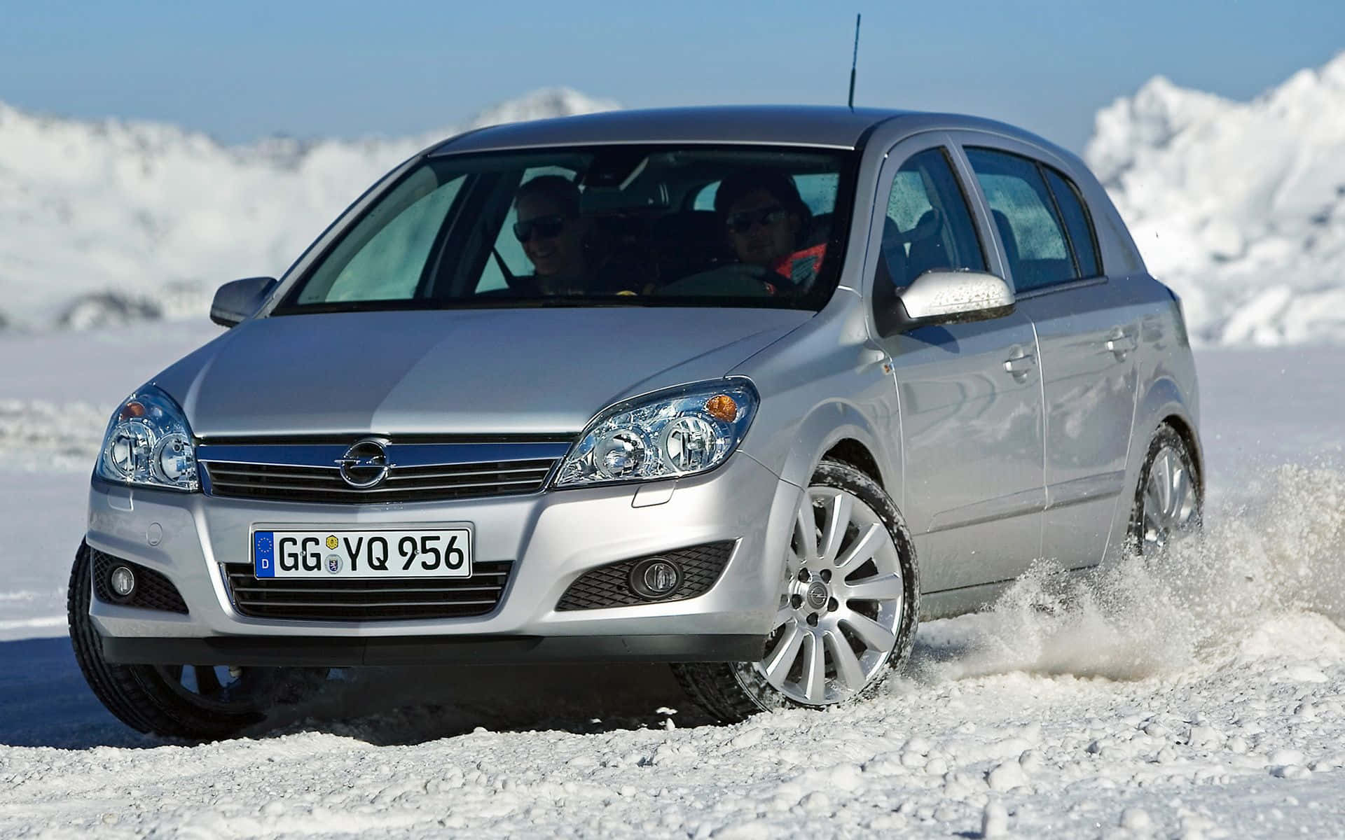 Opel Astra 桌布 壁紙 — 一輛銀色歐寶雅特在深雪中行駛，車內有兩個人，背景是白雪覆蓋的山脈。