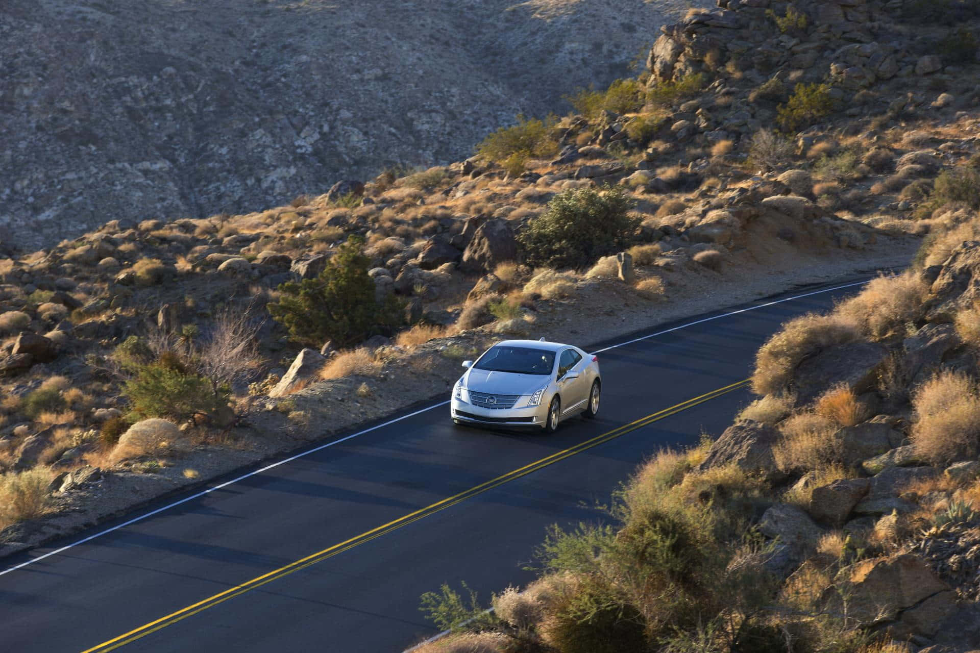 Cadillac Elr 桌布 壁紙 — 一輛銀色汽車在蜿蜒的沙漠公路上行駛，兩旁是岩石地形和乾燥灌木，沐浴在溫暖的陽光下。