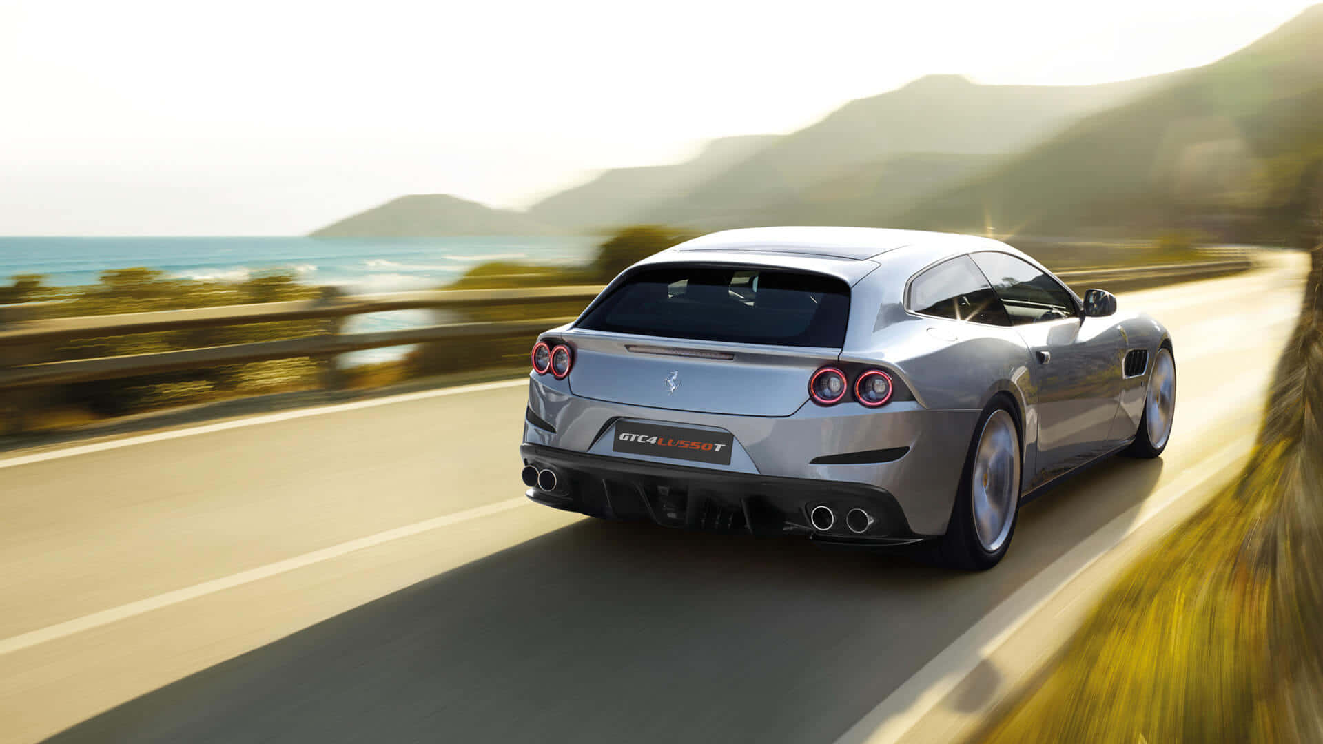 Ferrari Gtc4lusso 桌布 壁紙 — 一輛銀色法拉利 GTC4Lusso T 在陽光照耀的山巒和海洋背景下，沿著海岸高速公路疾馳，呈現模糊的動態效果。