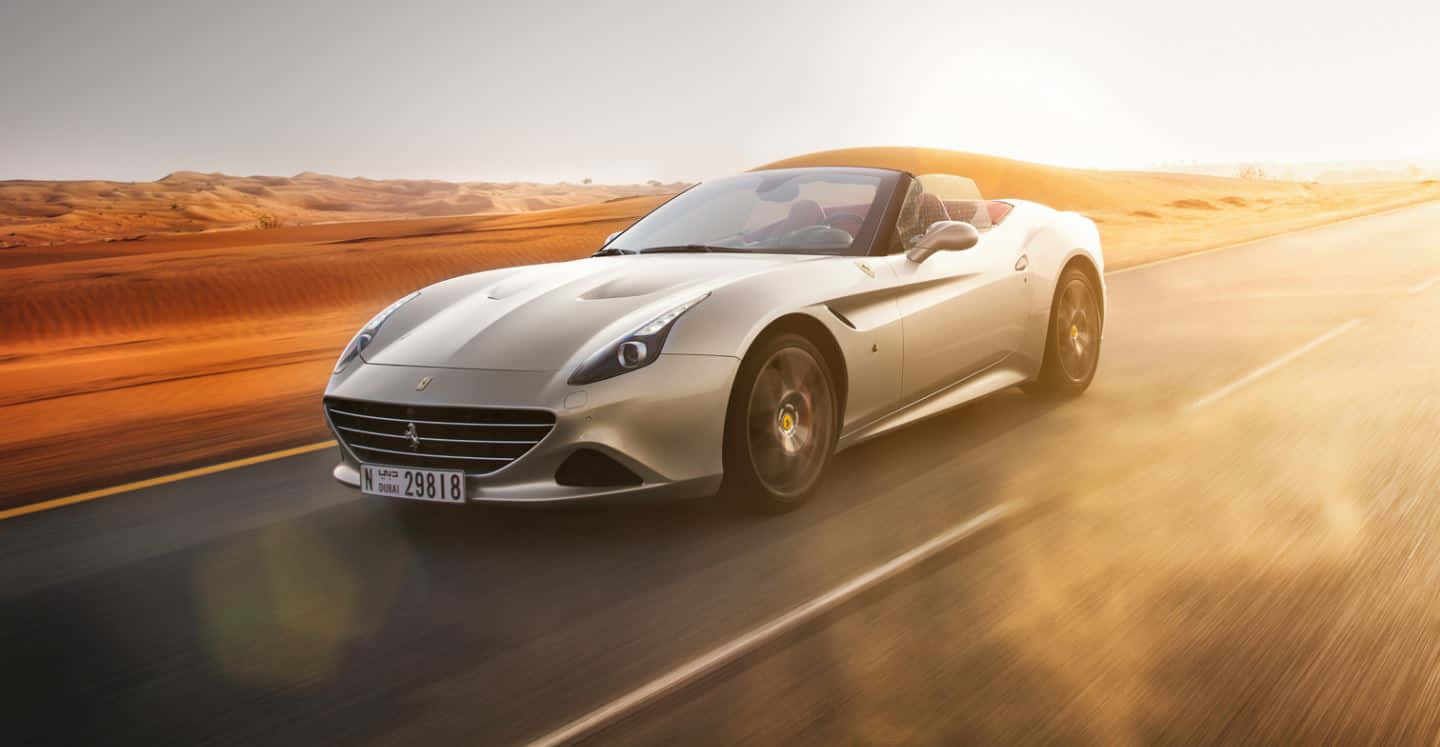 Ferrari California T 桌布 壁紙 — 一輛銀色法拉利開篷跑車在日落時分沿沙漠公路疾馳，在沙丘和模糊的動態上投下金色光芒。